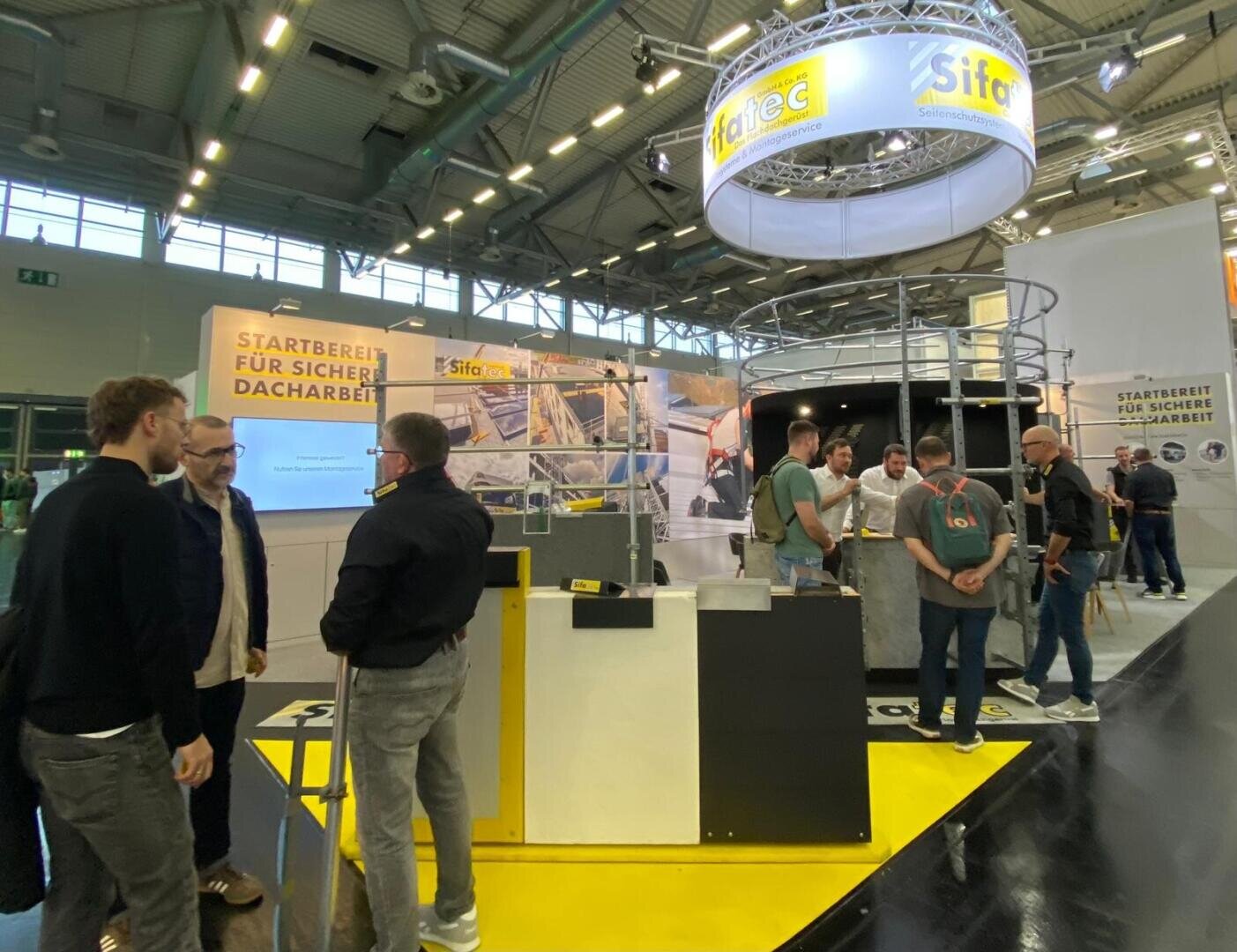 In einer geräumigen, gut beleuchteten Messehalle mit hohen Decken und Industriebeleuchtung versammeln sich Menschen und unterhalten sich an einem Messestand mit Sicherheitsausrüstungen und großen Schildern in deutscher Sprache.