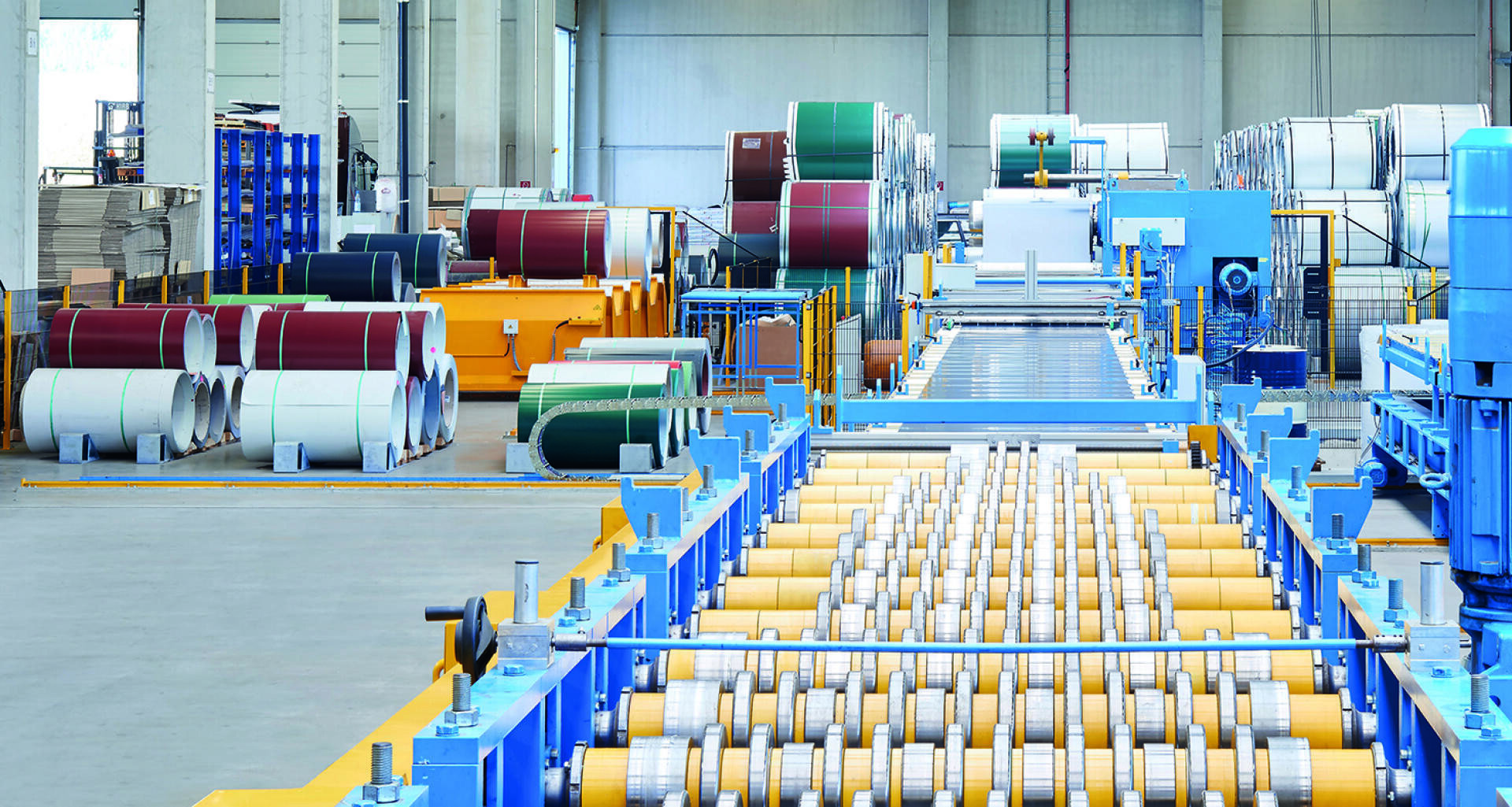 Ein modernes Fabrikinterieur mit großen Metallspulen, Industriemaschinen und Förderrollen für die Blechverarbeitung; verschiedenfarbige Spulen und Geräte sind in der geräumigen, gut beleuchteten Anlage angeordnet.