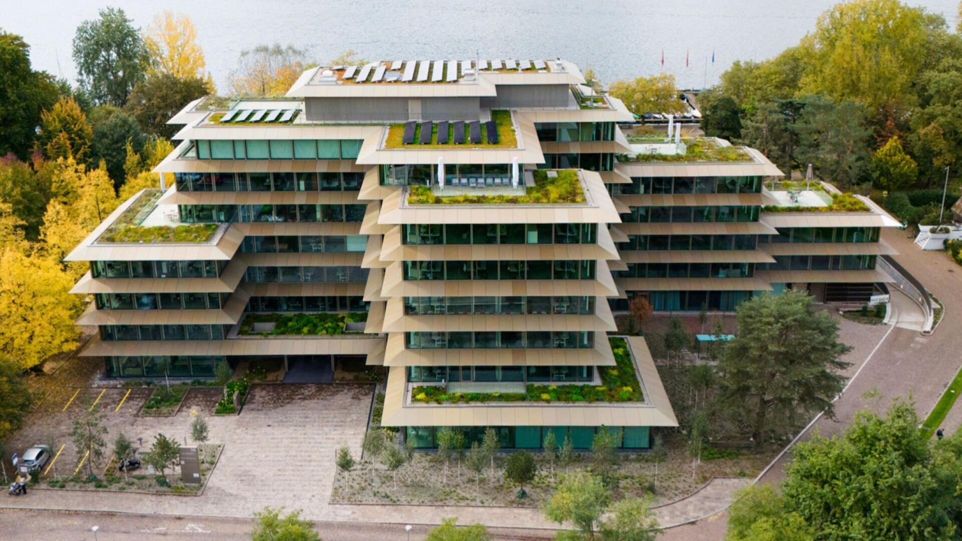 A modern, multi-storey building with wide, stepped and green balconies. The surrounding trees are colored in autumnal hues and a body of water can be seen behind the building. The architecture is characterized by glass walls and flat roofs.