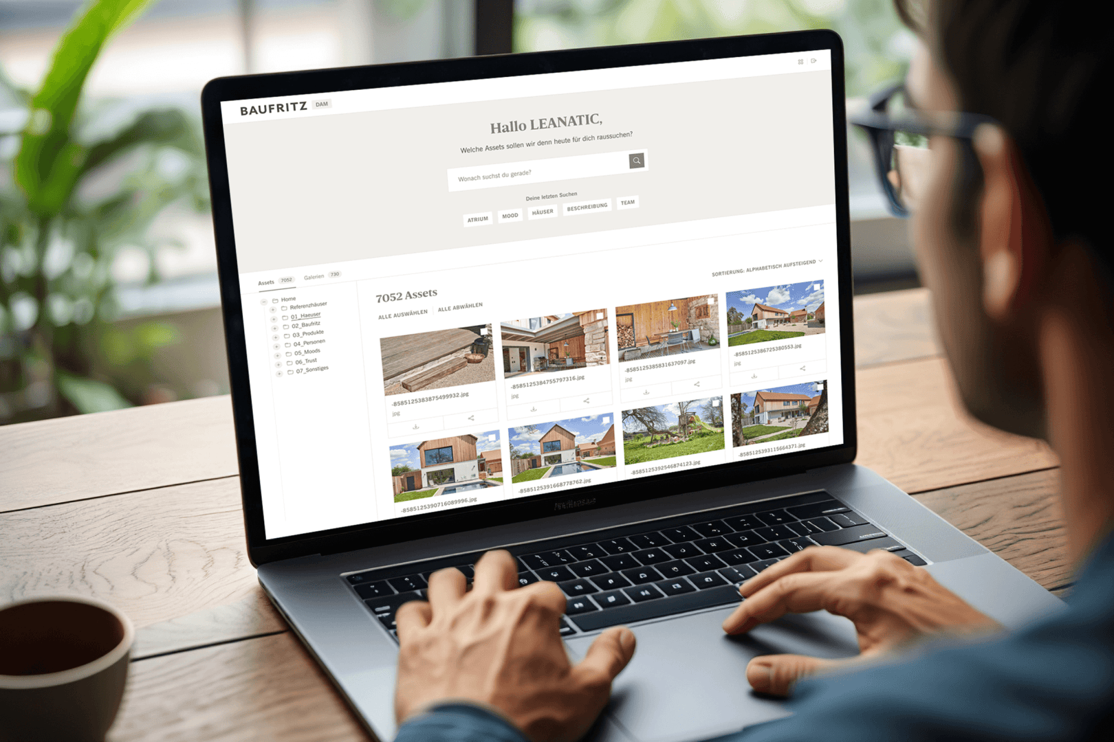 Eine Person benutzt einen Laptop, auf dem eine Website mit Immobilienbildern und -angeboten angezeigt wird, und durchsucht die mit Hausfotos, Beschreibungen und Ordnern versehenen Objekte. In der Kopfzeile der Website steht "Hallo LEANATIC".