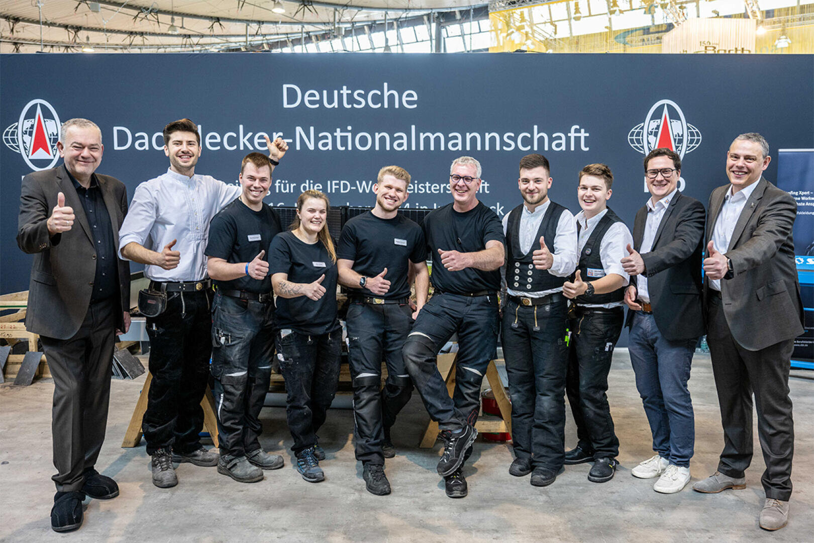 A group of ten people, some in work uniforms, others in business attire, stand smiling and with their thumbs up in front of a sign that reads German National Roofing Team.
