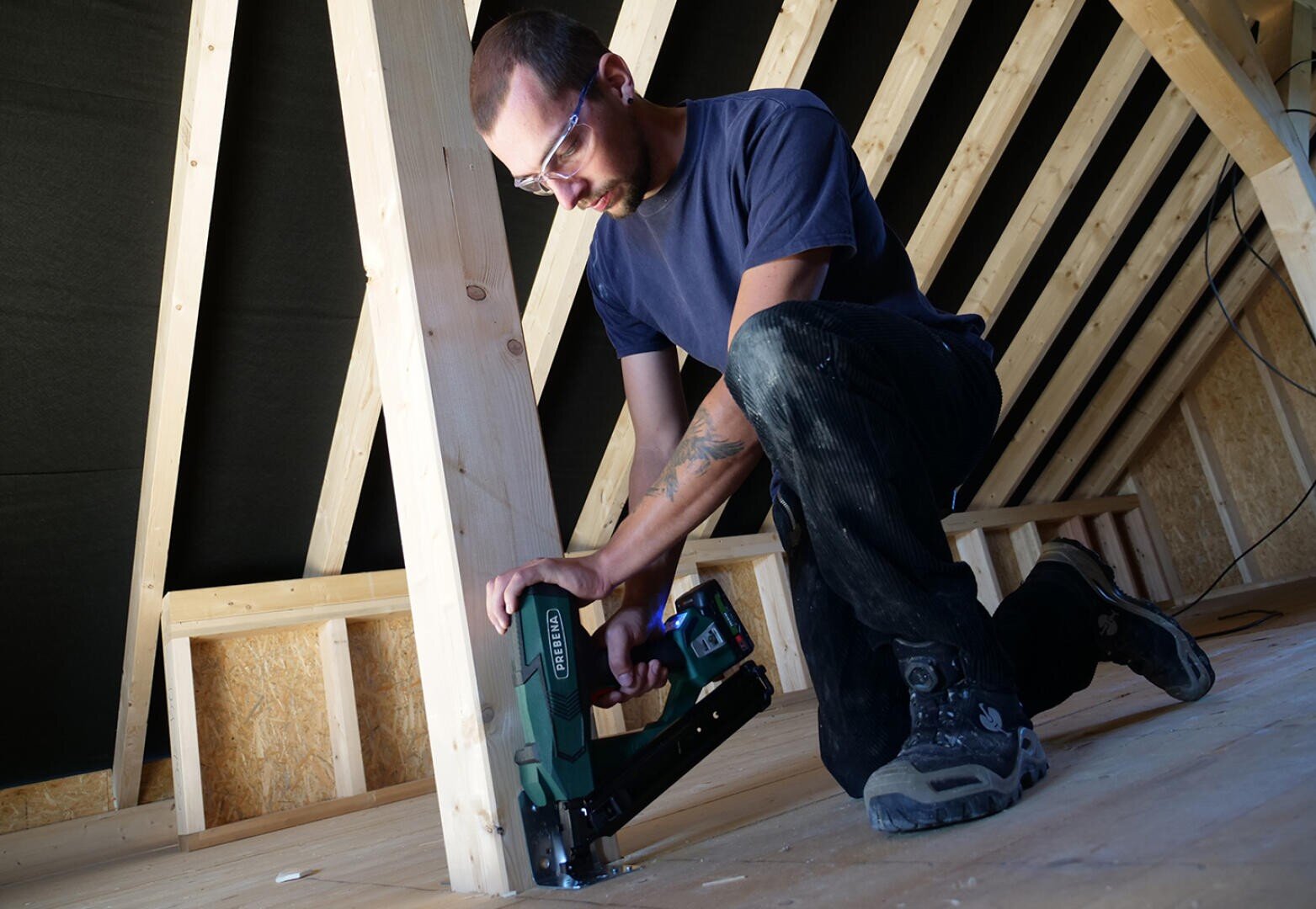 A person wearing safety glasses uses a green and black power tool to nail or staple flooring in a wooden attic under construction. The setting features exposed beams and unfinished wood.