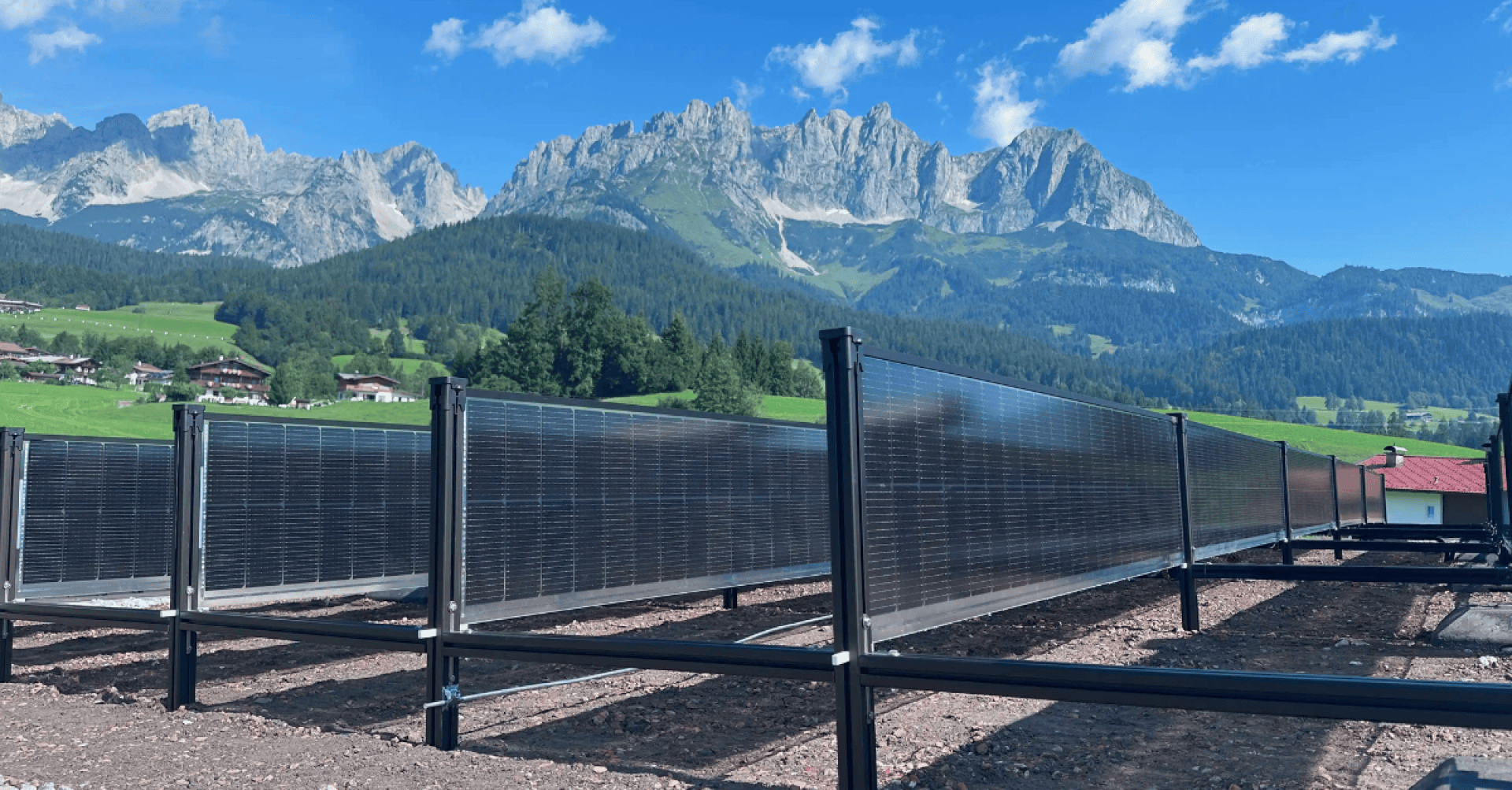 Rows of vertical solar modules stand on a gravel surface in front of a lush green landscape with houses, trees and high, rugged mountains under a bright blue sky with scattered clouds.