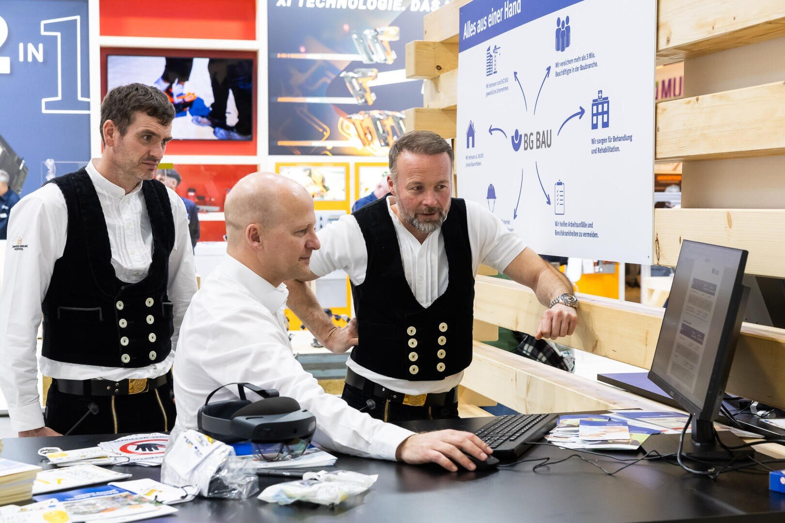 Drei Männer, zwei in traditioneller Tischlerkleidung und einer im weißen Hemd, stehen an einem Schreibtisch. Der Mann in Weiß bedient einen Computer, während die anderen zusehen. Im Hintergrund sind Holzbalken und Informationstafeln zu sehen.