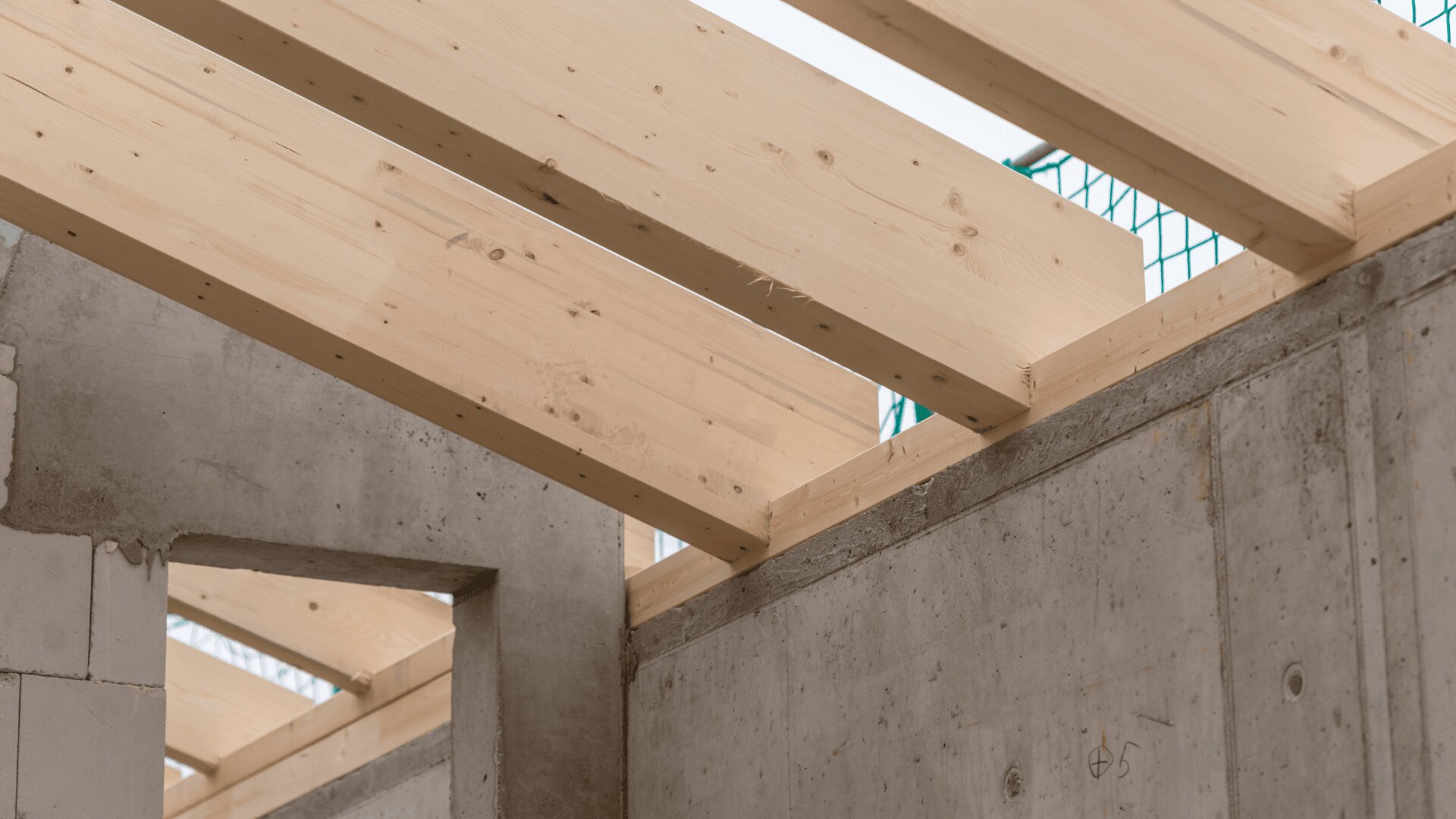 Wooden beams are installed on top of concrete walls, forming the framework of a building under construction. Natural light illuminates the unfinished structure.