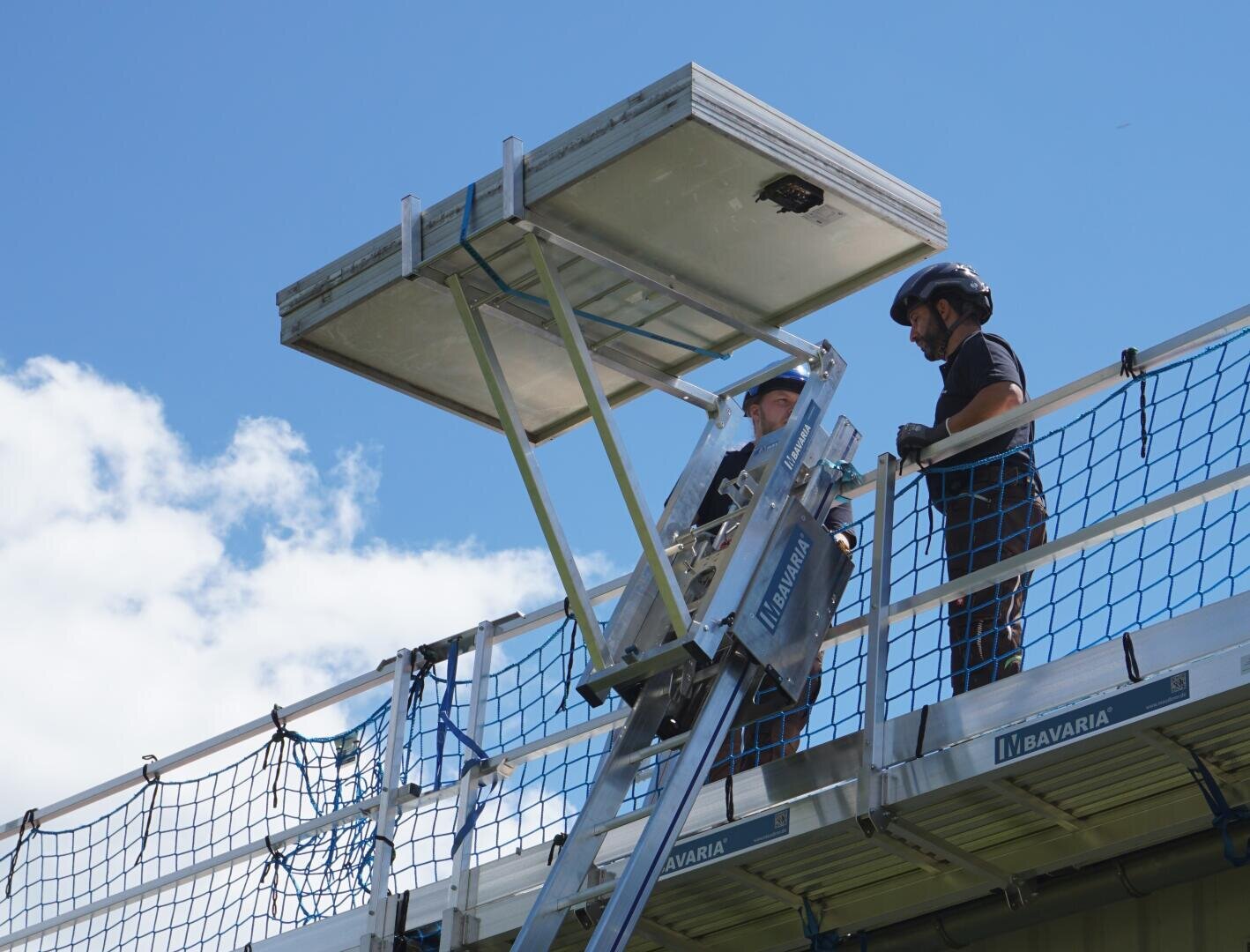 Zwei Personen in Schutzkleidung stehen auf einer Hebebühne und heben an einem klaren, sonnigen Tag ein großes Solarmodul auf ein Dach. Am Rand des Daches sind Sicherheitsnetze und Geländer zu sehen.