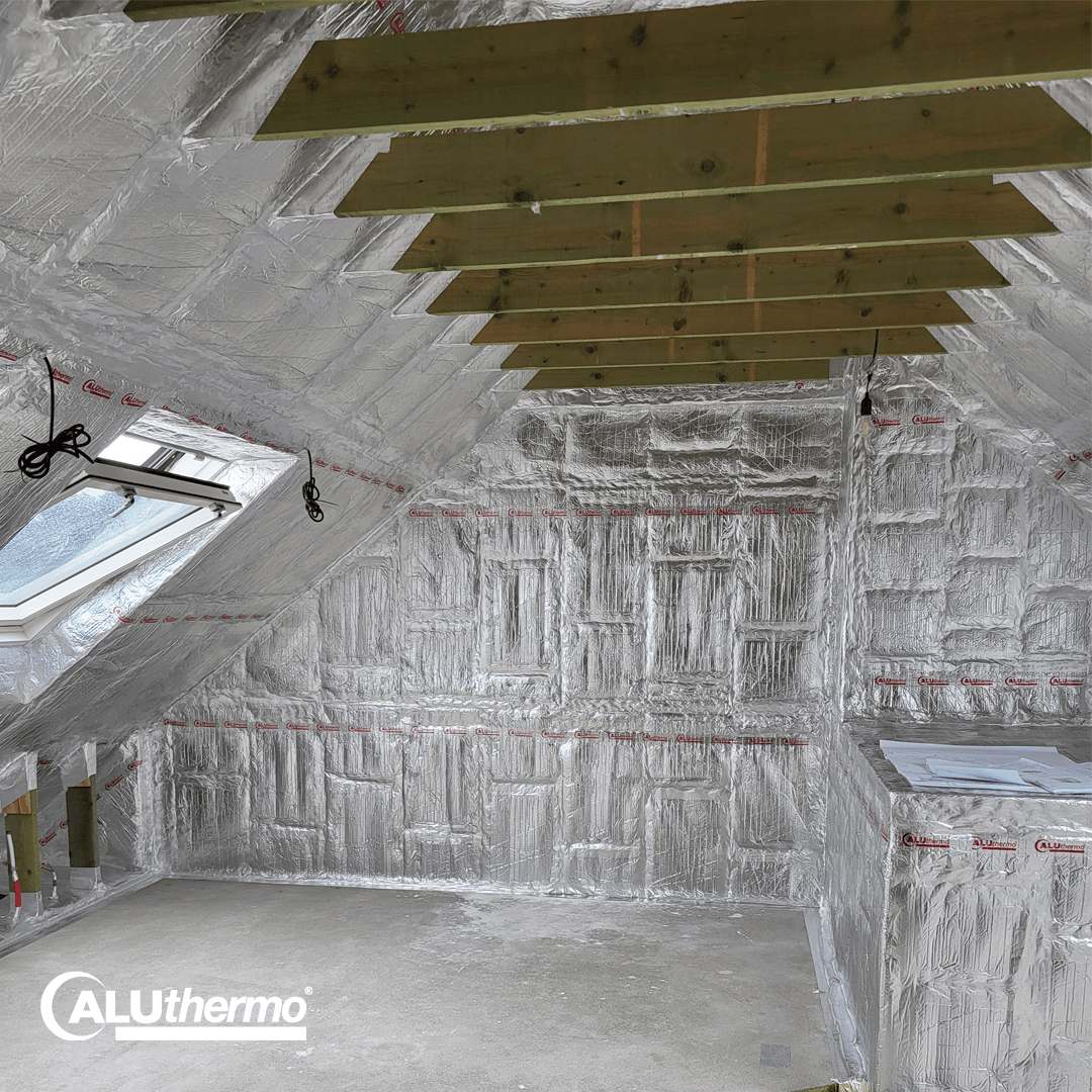 An attic under renovation with silver insulation material on the walls and ceiling, wooden beams on the ceiling, a skylight and the ALUthermo logo in the bottom left corner.