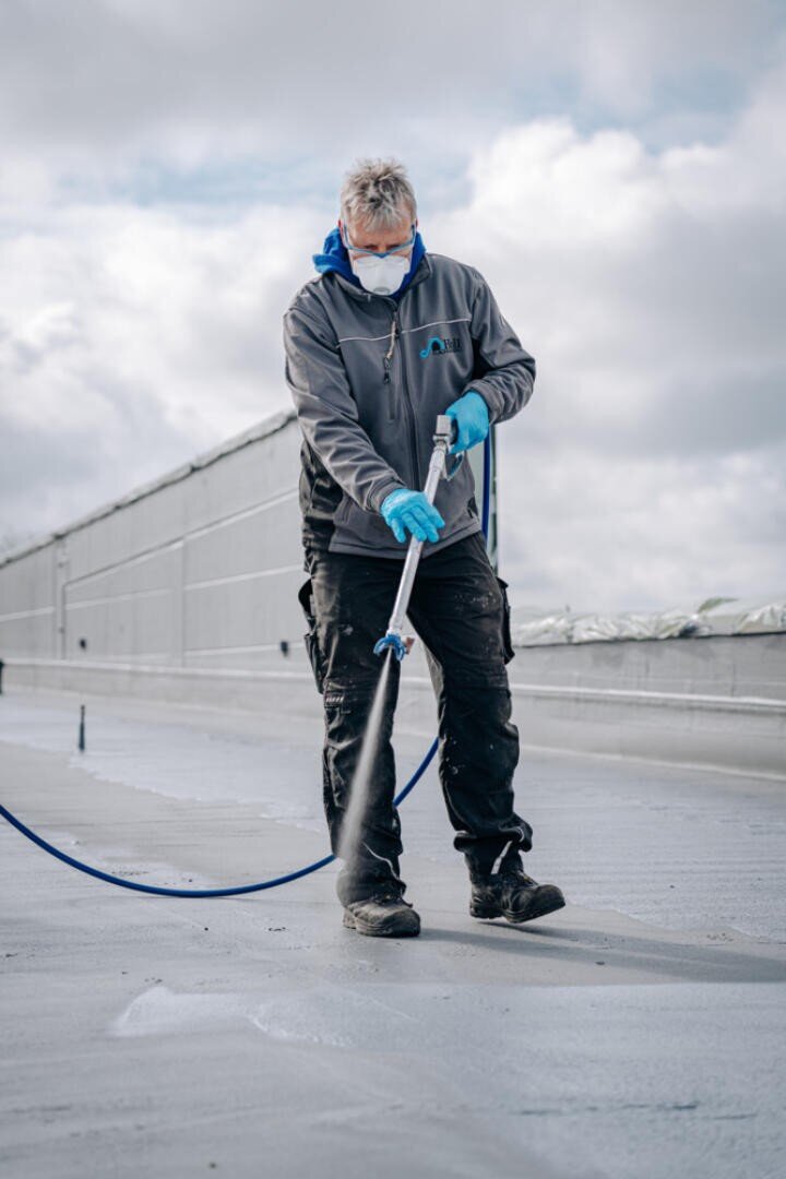Eine Person, die eine Gesichtsmaske, Handschuhe und eine graue Jacke trägt, sprüht mit einem Schlauch eine Flüssigkeit auf eine flache Dachfläche, während im Hintergrund der Himmel bewölkt ist.