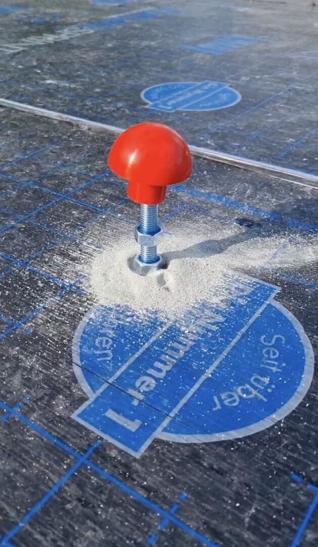 Ein großer Metallbolzen mit einer roten pilzförmigen Kappe ist in eine blau markierte Bauplatte geschraubt, die von weißem Pulver oder Staub umgeben ist.