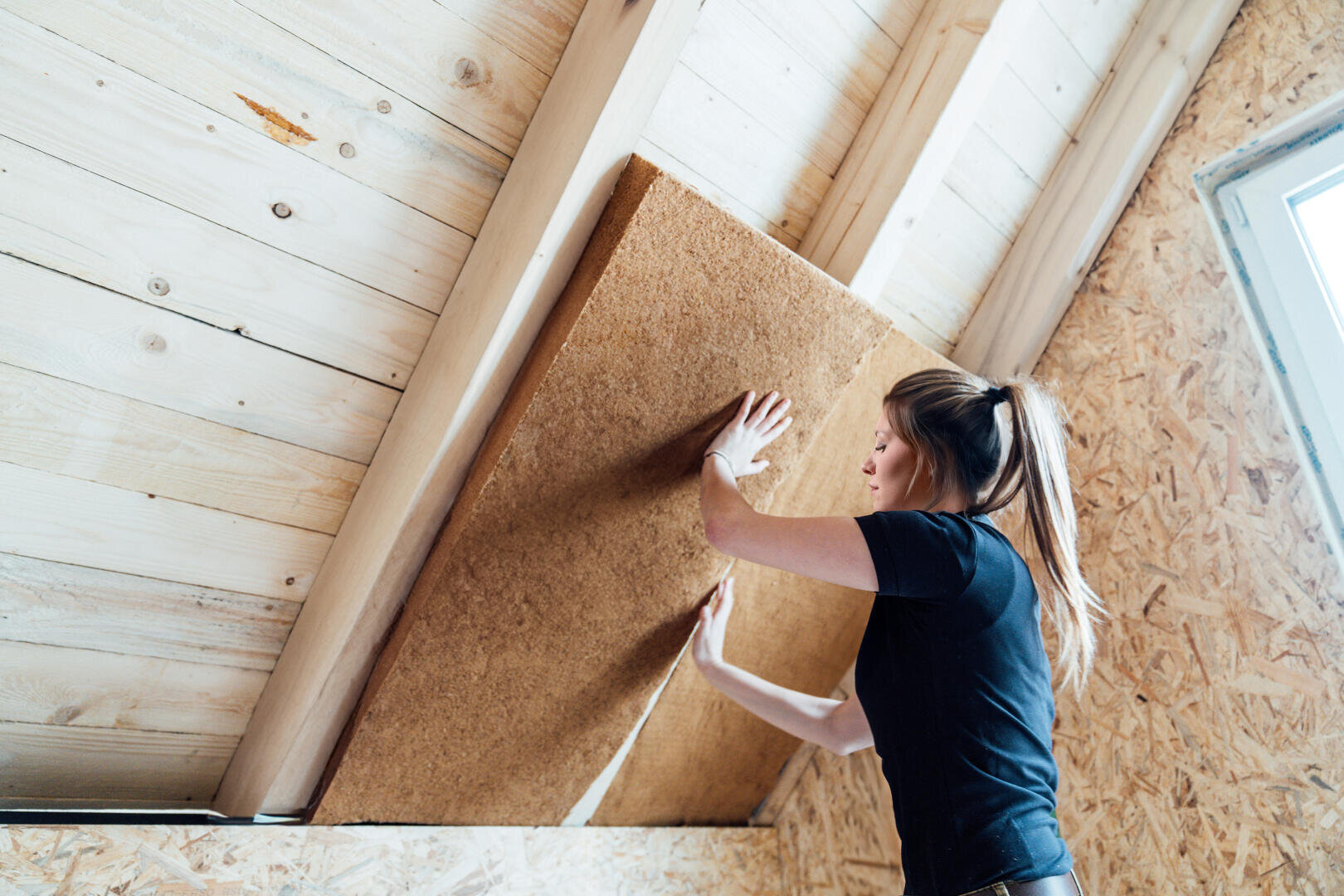Eine Frau in einem schwarzen Hemd bringt eine große Dämmplatte unter dem Holzdach eines Dachbodens an, wobei natürliches Licht aus einem nahe gelegenen Fenster einfällt.