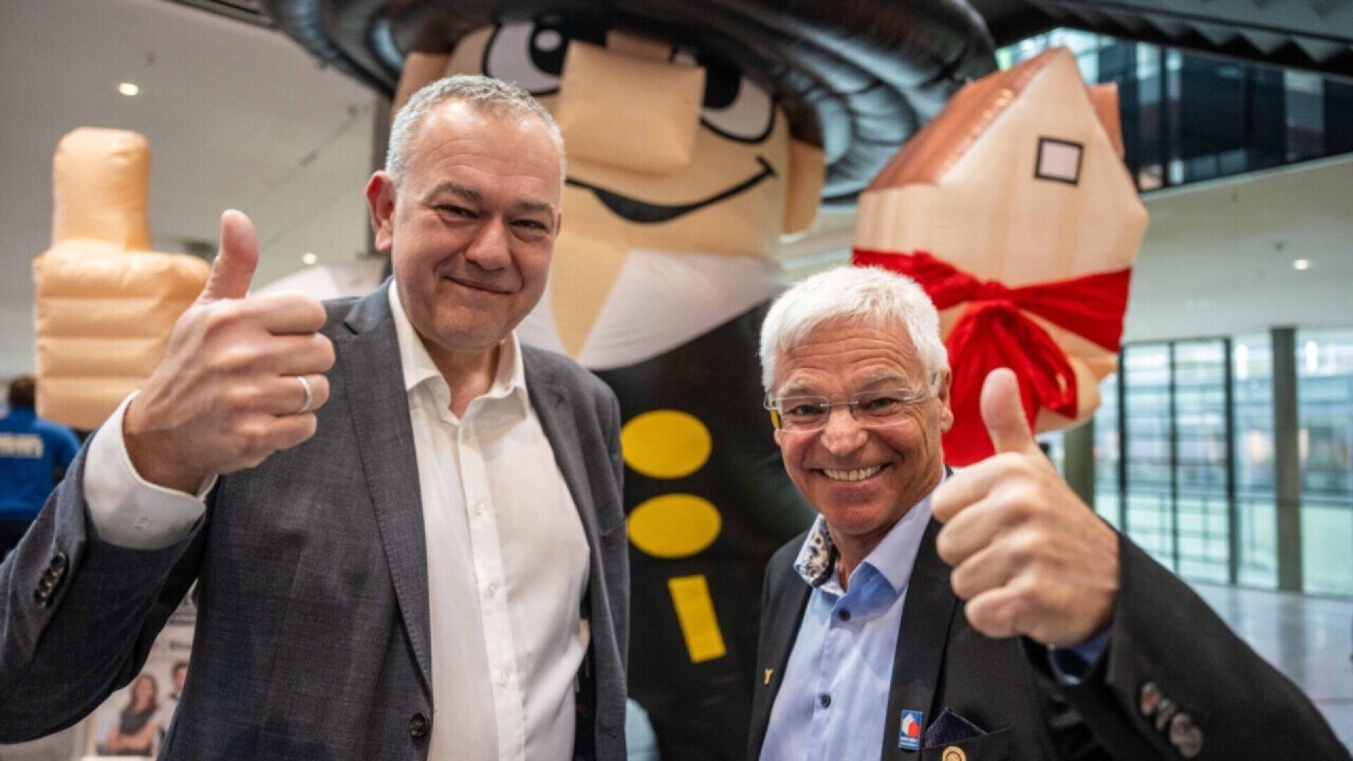 Two smiling men in suits give thumbs up in front of a large inflatable figure of a chimney sweep holding a house with a red bow, inside a modern building.