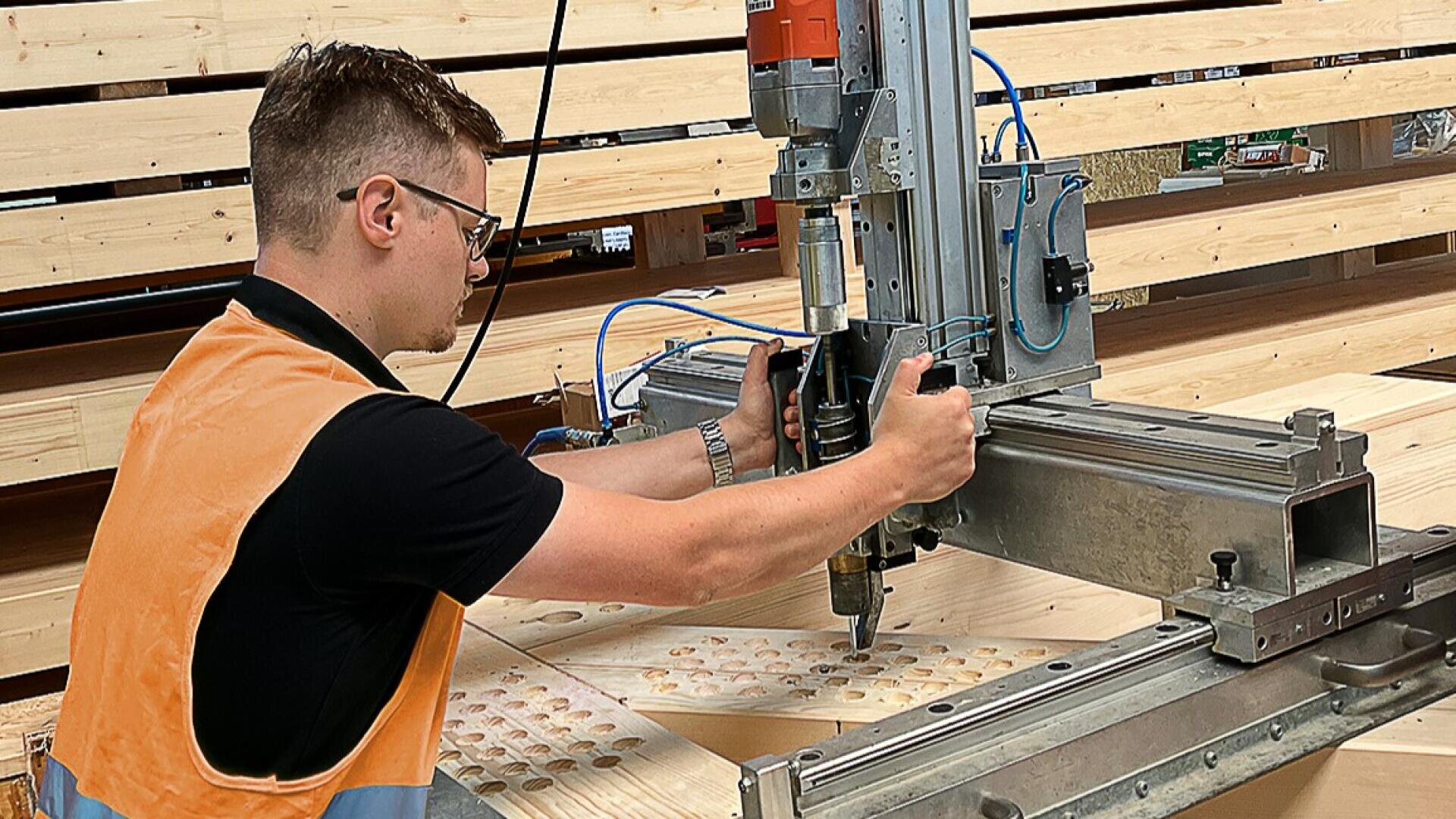 Eine Person, die eine Schutzbrille und eine orangefarbene Weste trägt, bedient eine große Industriemaschine, um Löcher in eine Holzplatte zu bohren, mit gestapelten Holzbrettern im Hintergrund.