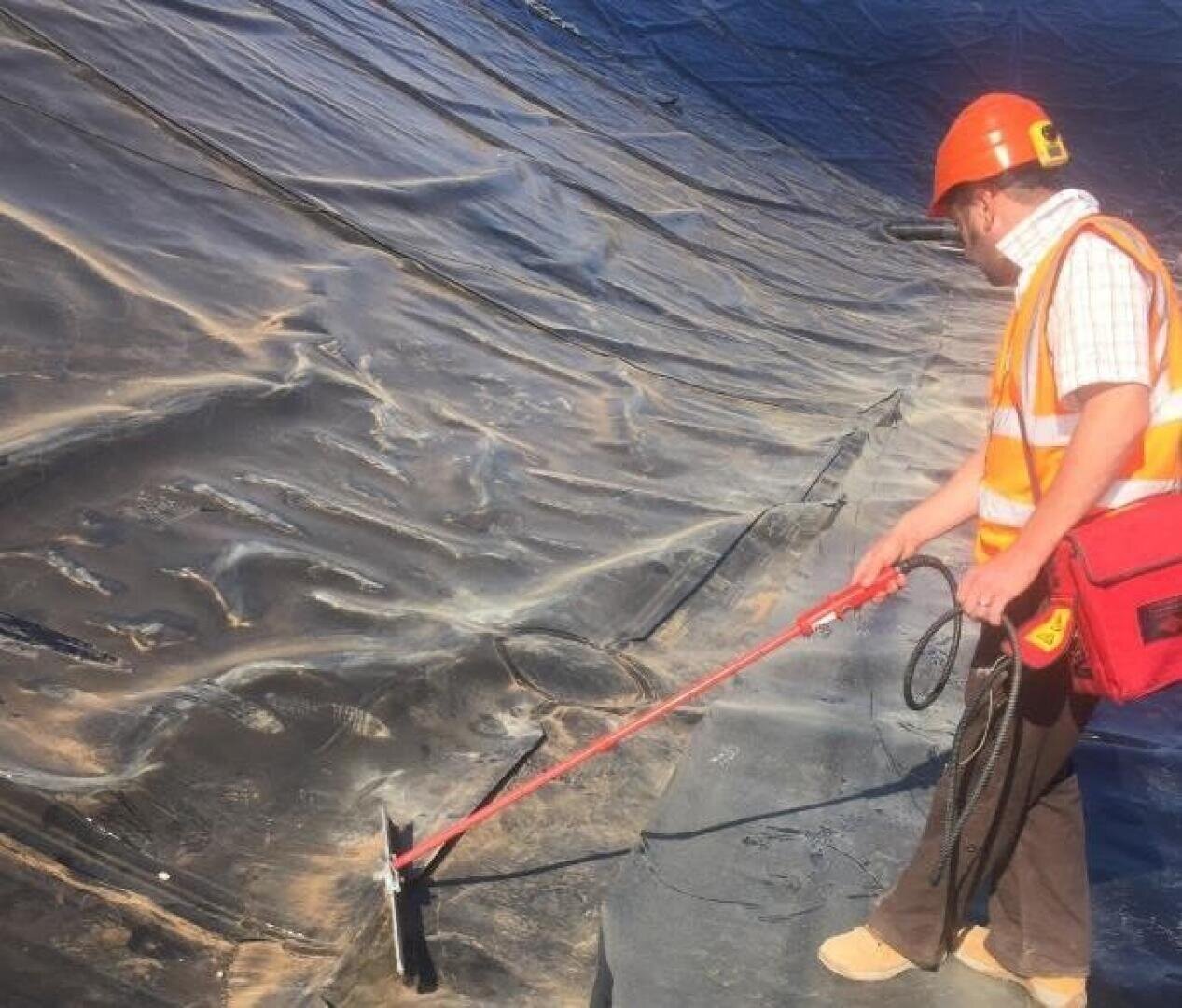 Ein Arbeiter mit Schutzhelm und Sicherheitsweste benutzt ein Werkzeug, um eine große schwarze Kunststofffolie, die eine Oberfläche bedeckt, zu inspizieren oder zu testen, möglicherweise auf einer Baustelle oder in einem Umweltbetrieb.