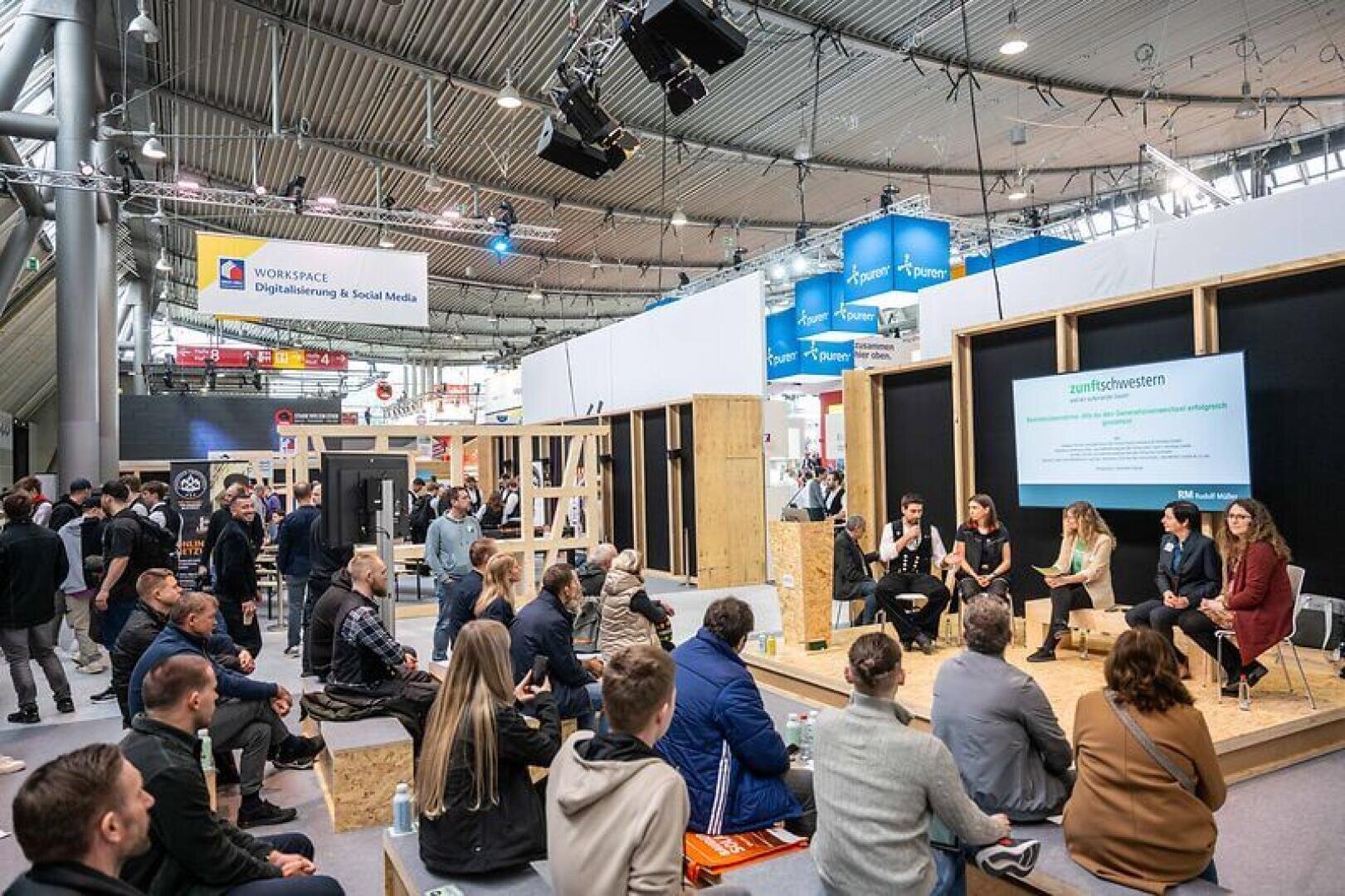 A group of people sit on benches and on the floor and listen to a panel discussion at an indoor event with modern displays and digital workplace signage in the background.