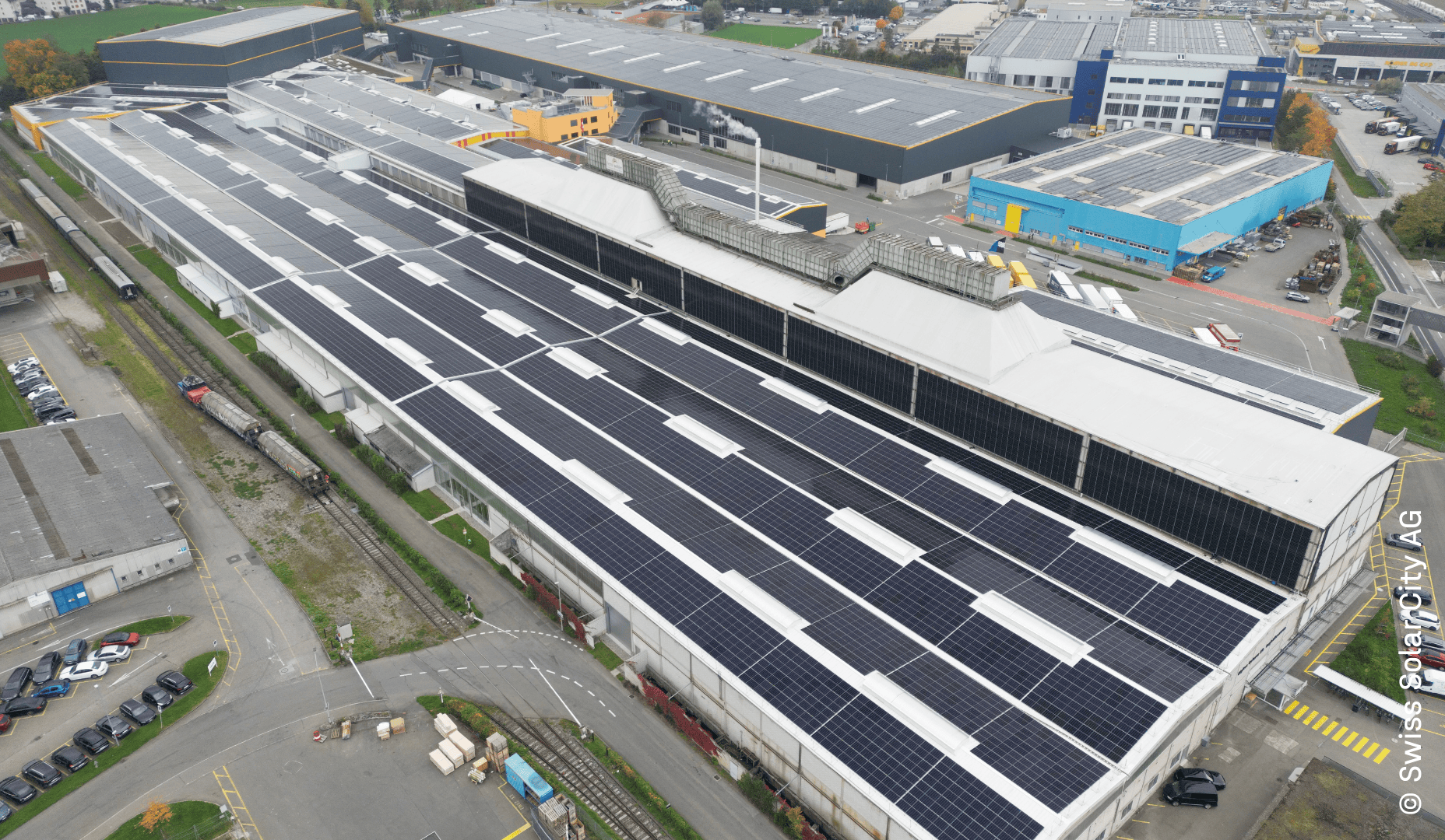 Ein großes Industriegebäude mit einem mit Solarzellen bedeckten Dach, umgeben von Straßen, geparkten Autos und anderen industriellen Strukturen in einem städtischen Gebiet.