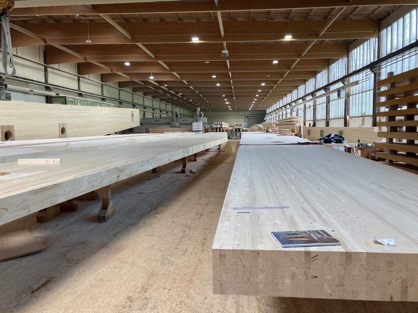 Große Holzbalken und -platten sind in einer geräumigen, gut beleuchteten Werkstatt mit hohen Decken und großen Fenstern an einer Seite angeordnet. In dem organisierten Arbeitsbereich sind verschiedene Werkzeuge und Materialien zu sehen.