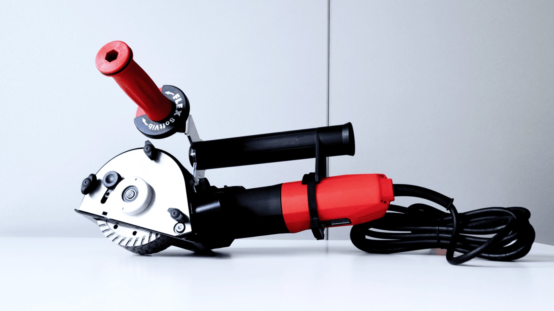Ein rot-schwarzer elektrischer Winkelschleifer mit Kabel und Seitengriff liegt auf einer weißen Oberfläche vor einem hellgrauen Hintergrund.