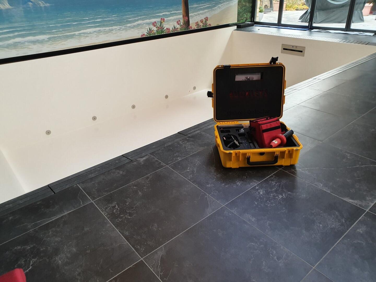 Ein gelber Hartschalenkoffer mit verschiedenen Werkzeugen und Ausrüstungsgegenständen steht offen auf einem dunklen Fliesenboden neben einem leeren Hallenbad mit einem Wandgemälde mit einer Strandszene an der Wand.