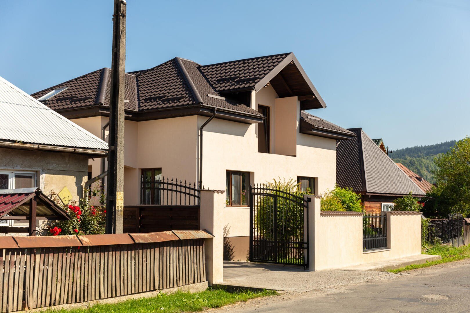 Ein modernes beigefarbenes Haus mit einem dunkelbraunen Dach und einem schwarzen Metalltor, umgeben von einer niedrigen cremefarbenen Mauer, neben einem älteren Holzzaun und anderen Häusern an einem sonnigen Tag. Im Hintergrund sind Bäume und Hügel zu sehen.