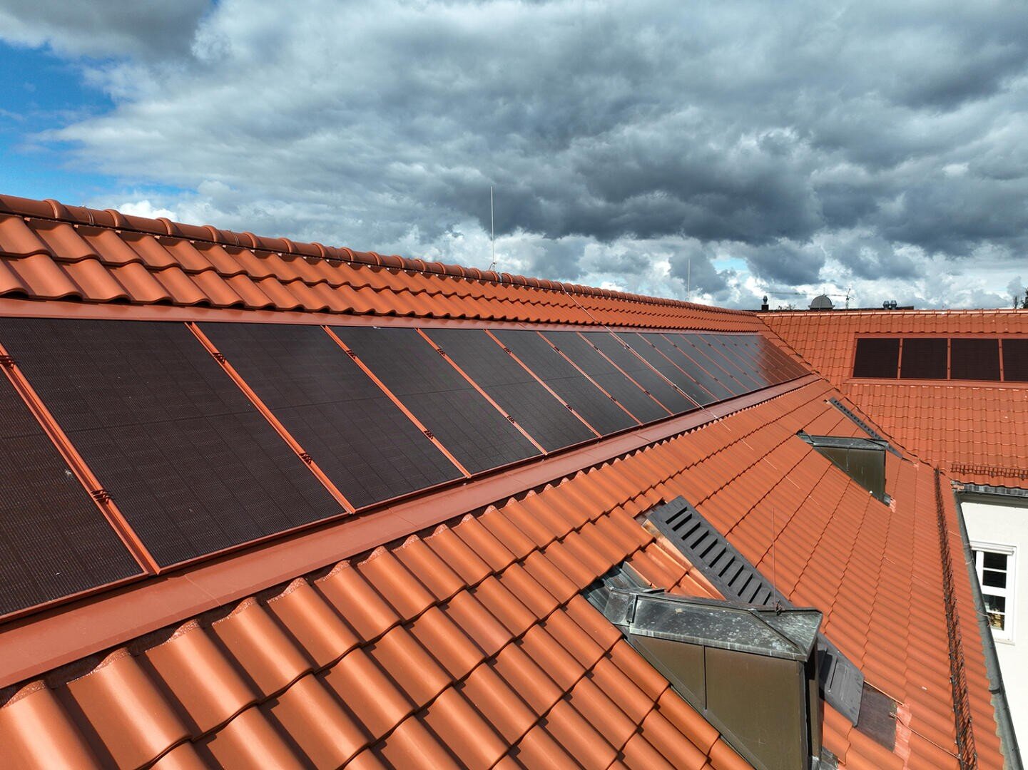 Auf einem orangefarbenen Ziegeldach installierte Solarmodule bei teilweise bewölktem Himmel, wobei das Sonnenlicht durch die Wolken bricht.