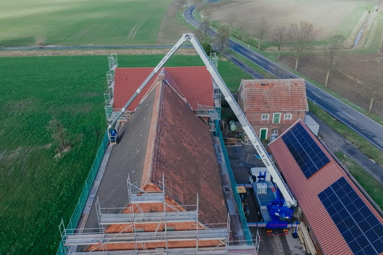 Aerial view of a building with a red tiled roof being renovated, surrounded by scaffolding. A crane truck is parked next to the building and solar cells can be seen on an adjacent roof. Fields and a road can be seen in the background.