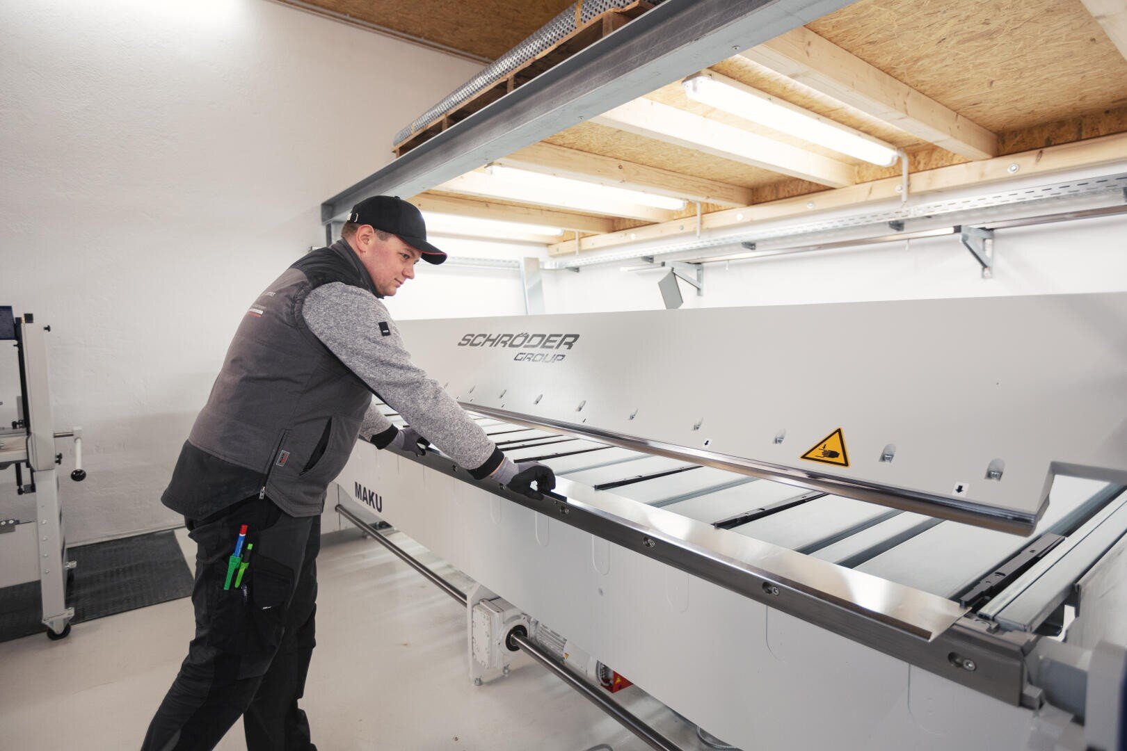 A man in work clothes and gloves operates a large industrial machine from the Schröder Group in a well-lit workshop. Tools can be seen in his bag. The room has white walls and a wooden ceiling.
