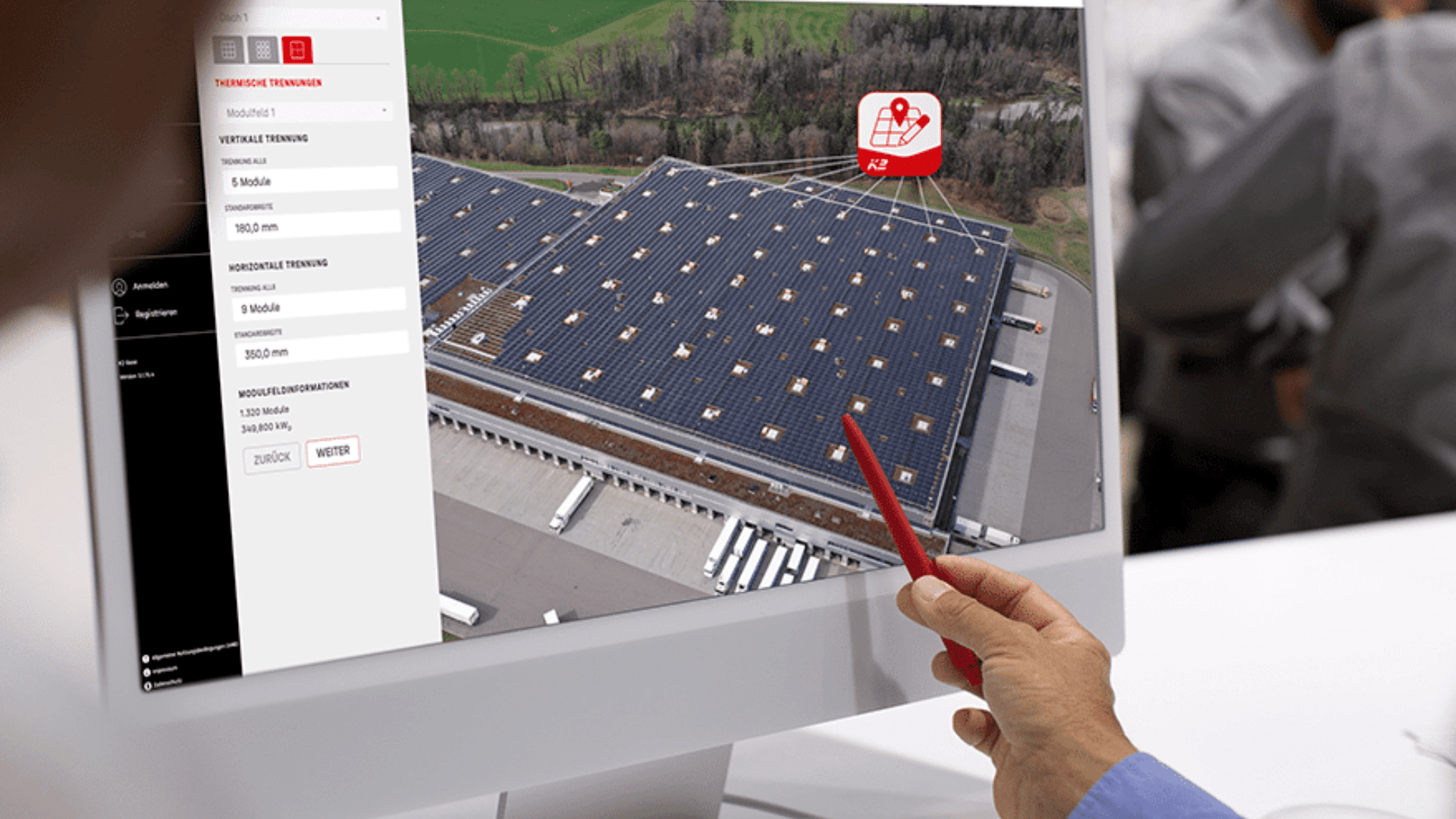 Eine Person zeigt mit einem roten Stift auf einen Computerbildschirm, auf dem Luftbilder eines großen Gebäudes mit Solarmodulen auf dem Dach und technischen Informationen auf der linken Seite des Bildschirms angezeigt werden.
