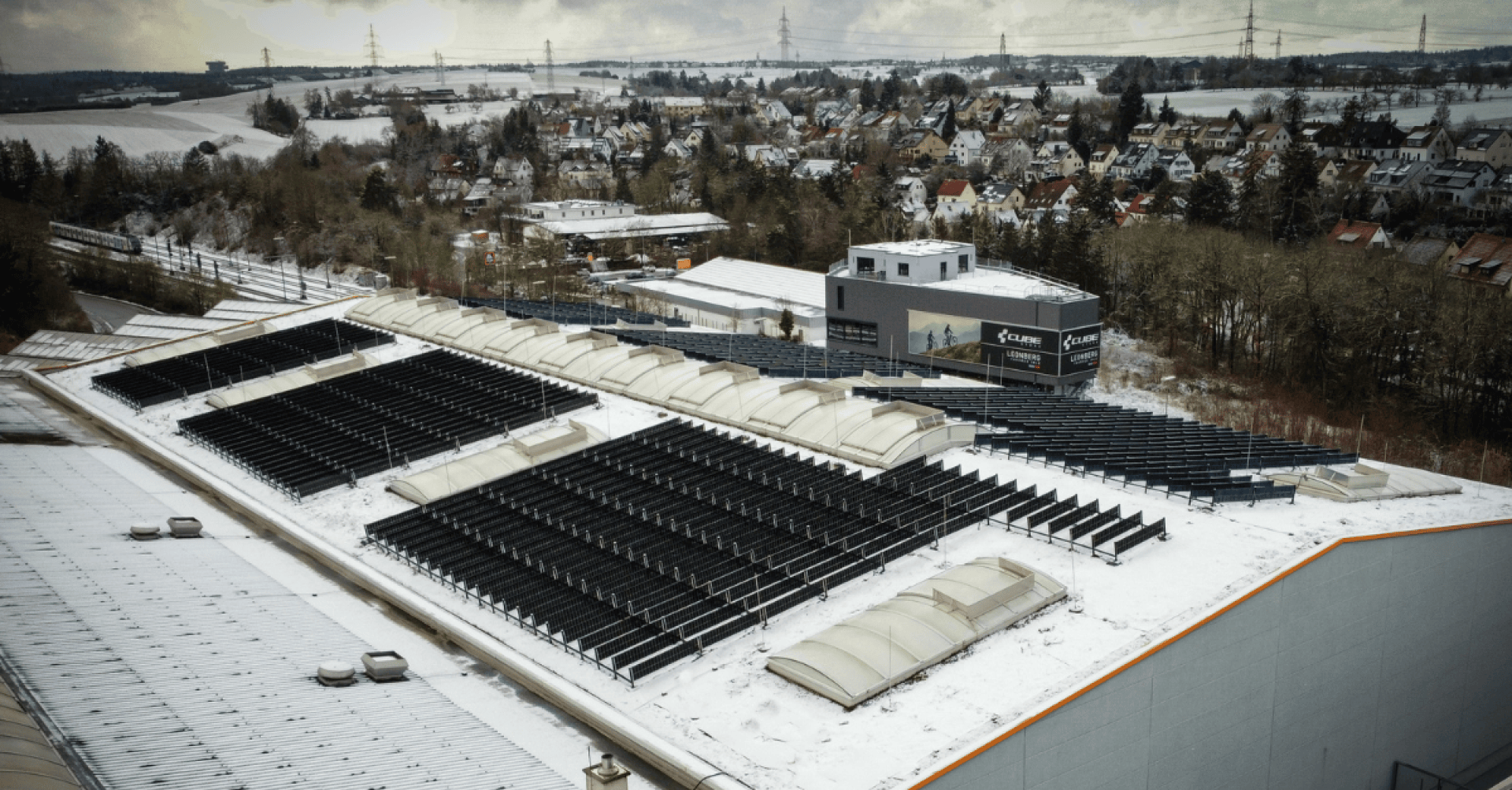 A large industrial building with snow-covered solar panels on the roof, surrounded by trees and a residential area in winter. The roofs and ground nearby are also covered in snow.