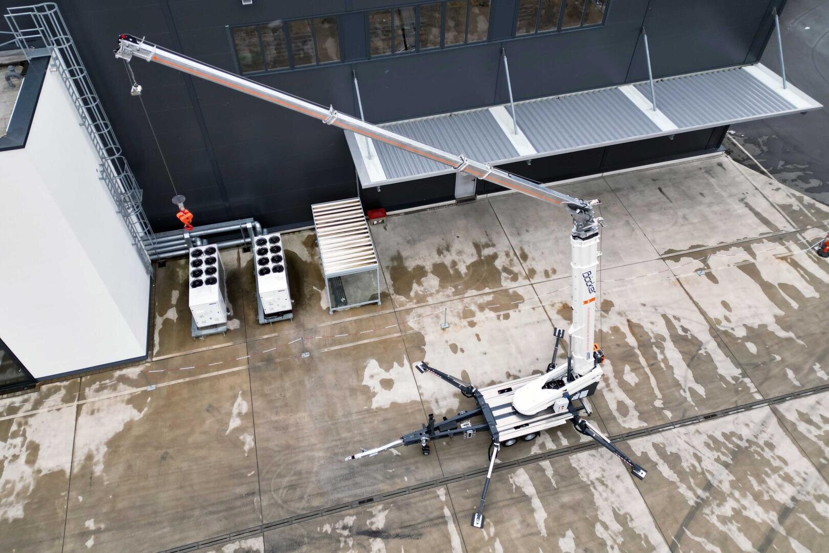 Aerial view of a white mobile crane with extended arm and stabilizing legs set up on a concrete surface in front of an industrial building. Various pipes and roof structures can be seen nearby.