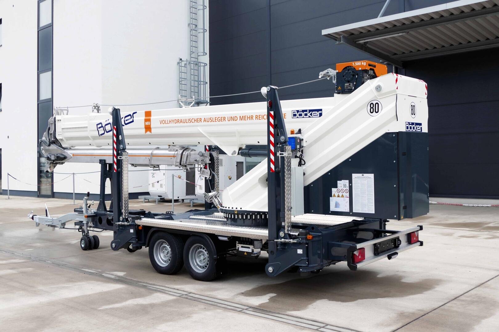 A white mobile crane with extendable jib arms is parked in front of an industrial building. The vehicle has two axles and support legs, the lettering Bocker can be seen on the side.