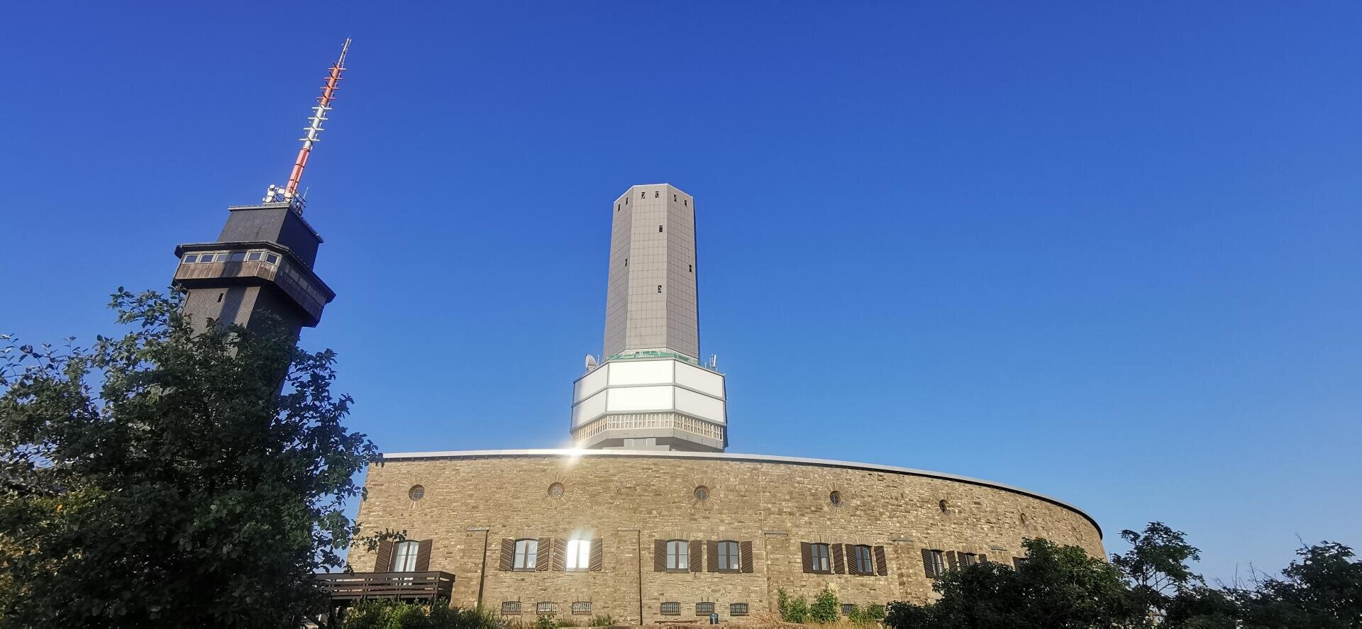 Ein rundes Steingebäude mit kleinen Fenstern und einem hohen achteckigen Turm in der Mitte, daneben ein schwarzer Funkmast, vor einem klaren blauen Himmel und umgeben von Grün.