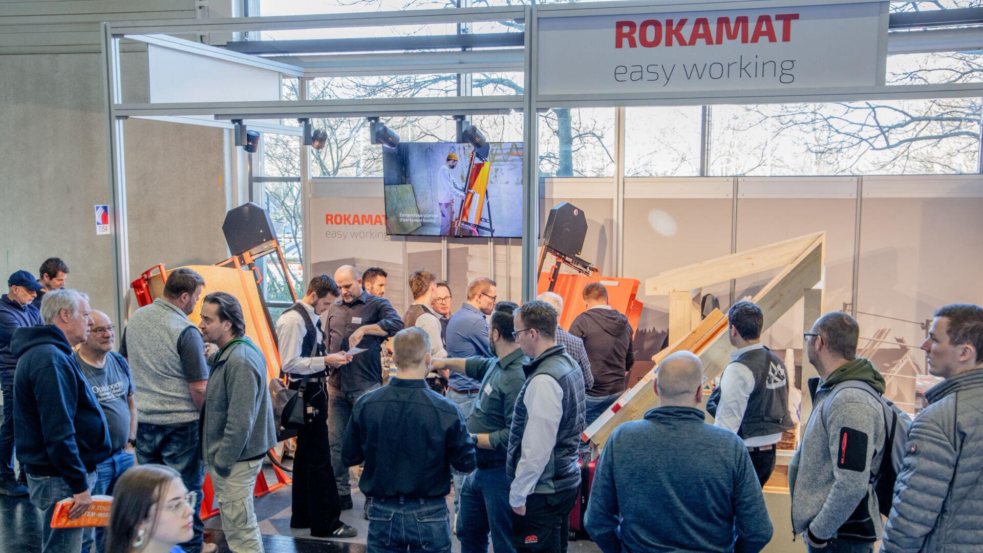 Ein überfüllter Messestand mit ROKAMAT easy working signage, auf dem die Besucher interagieren, Vorführungen beobachten und die ausgestellten Bauwerkzeuge und -geräte bei heller Innenbeleuchtung betrachten.