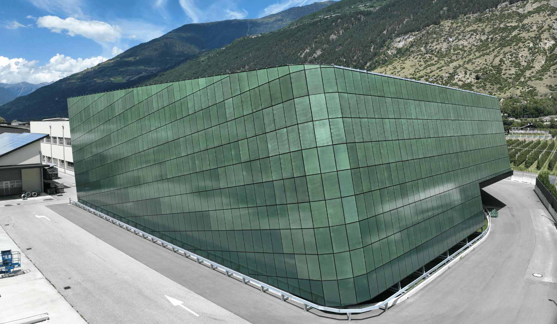 Ein großes, modernes, grünes Glasgebäude mit kantigem Design steht neben einer geschwungenen Straße, umgeben von Bergen und Grünflächen unter einem blauen Himmel mit vereinzelten Wolken.