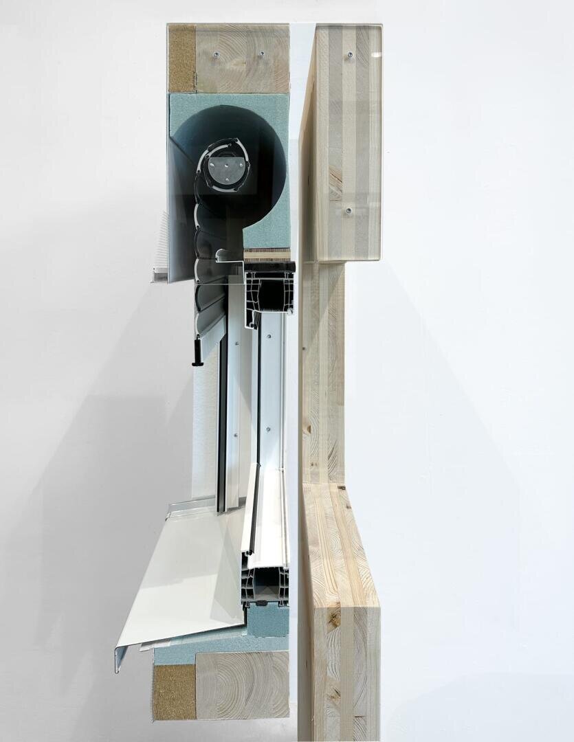 Querschnitt einer Wand mit Materialschichten, darunter Holz, Dämmung und Metallteile, mit einem aufgerollten Fensterjalousiemechanismus und einer nach außen ragenden Fensterbank.