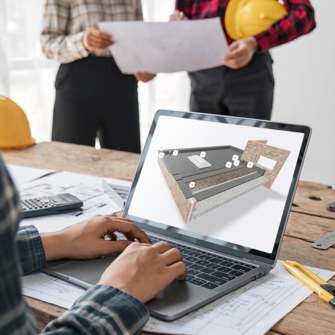 One person uses a laptop with a 3D building model on the screen, while two people in plaid shirts and hard hats examine construction plans on a wooden table in the background.