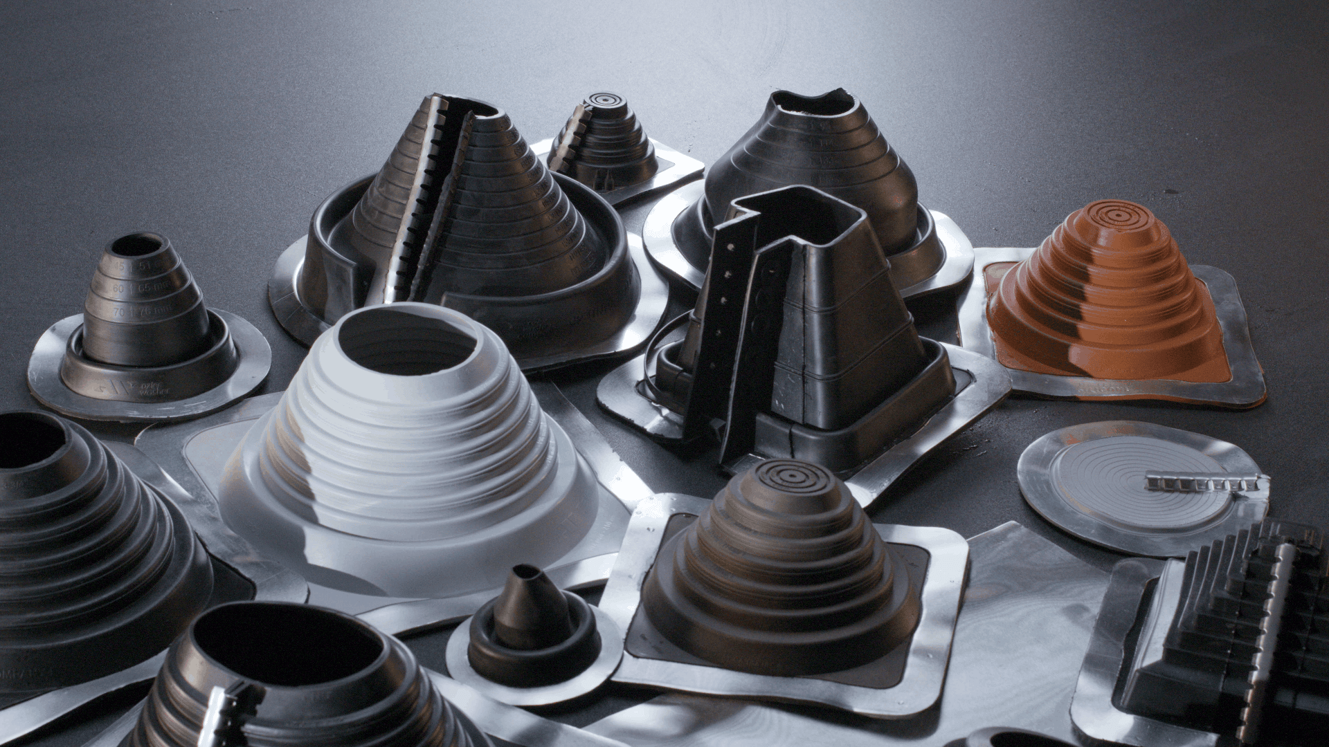 Eine Vielzahl von konischen und zylindrischen Dachabdeckungen aus Gummi in schwarzer, grauer und brauner Farbe sind auf einer Metalloberfläche angeordnet. Die Abdeckungen haben unterschiedliche Größen und Formen, mit gerippten Strukturen und quadratischen Grundflächen.