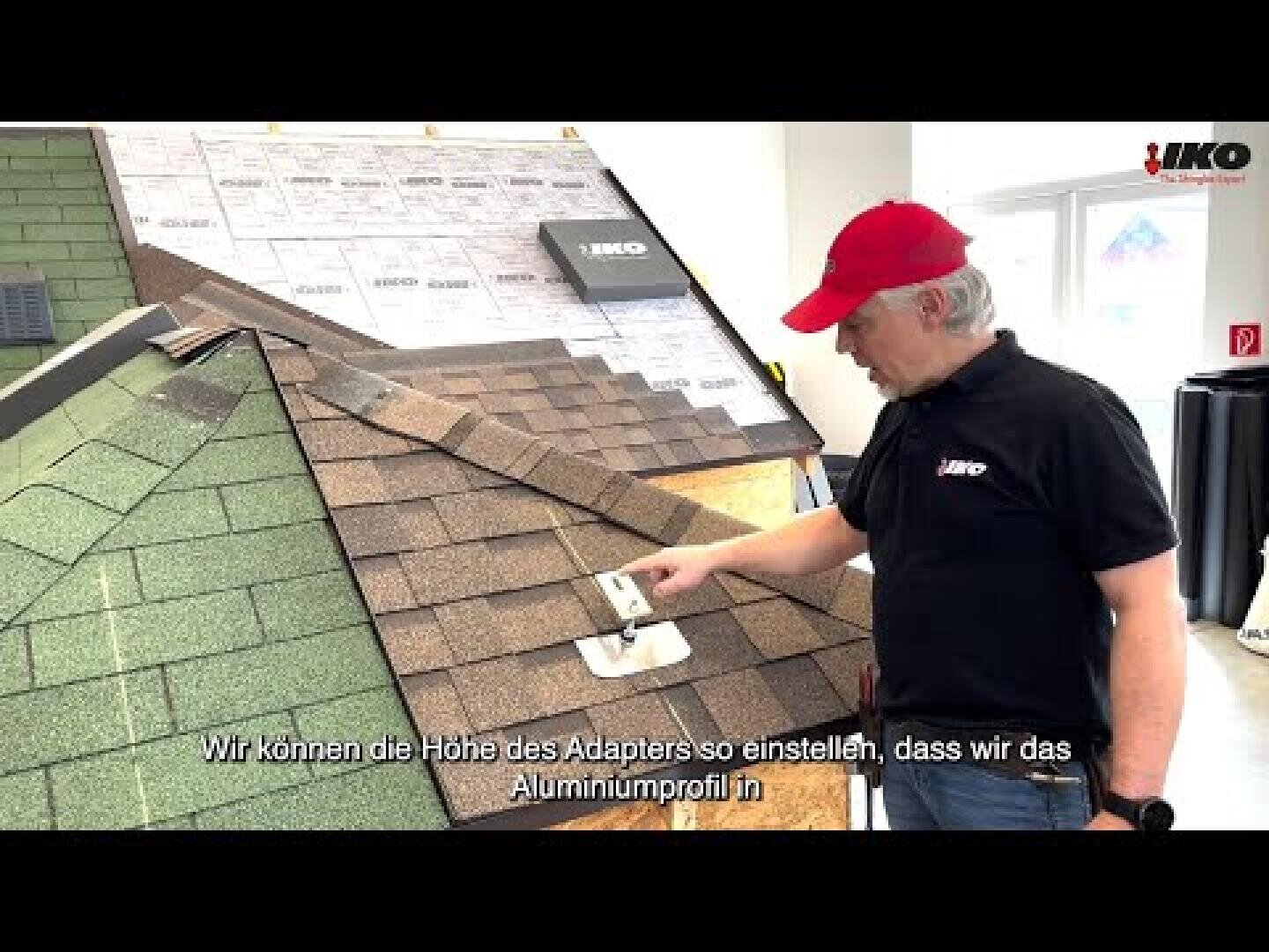 Ein Mann in schwarzem Hemd und roter Mütze demonstriert die Verlegung von Dachschindeln auf einem Modelldach in einem Ausstellungsraum. Am unteren Rand des Bildes werden deutsche Untertitel eingeblendet.