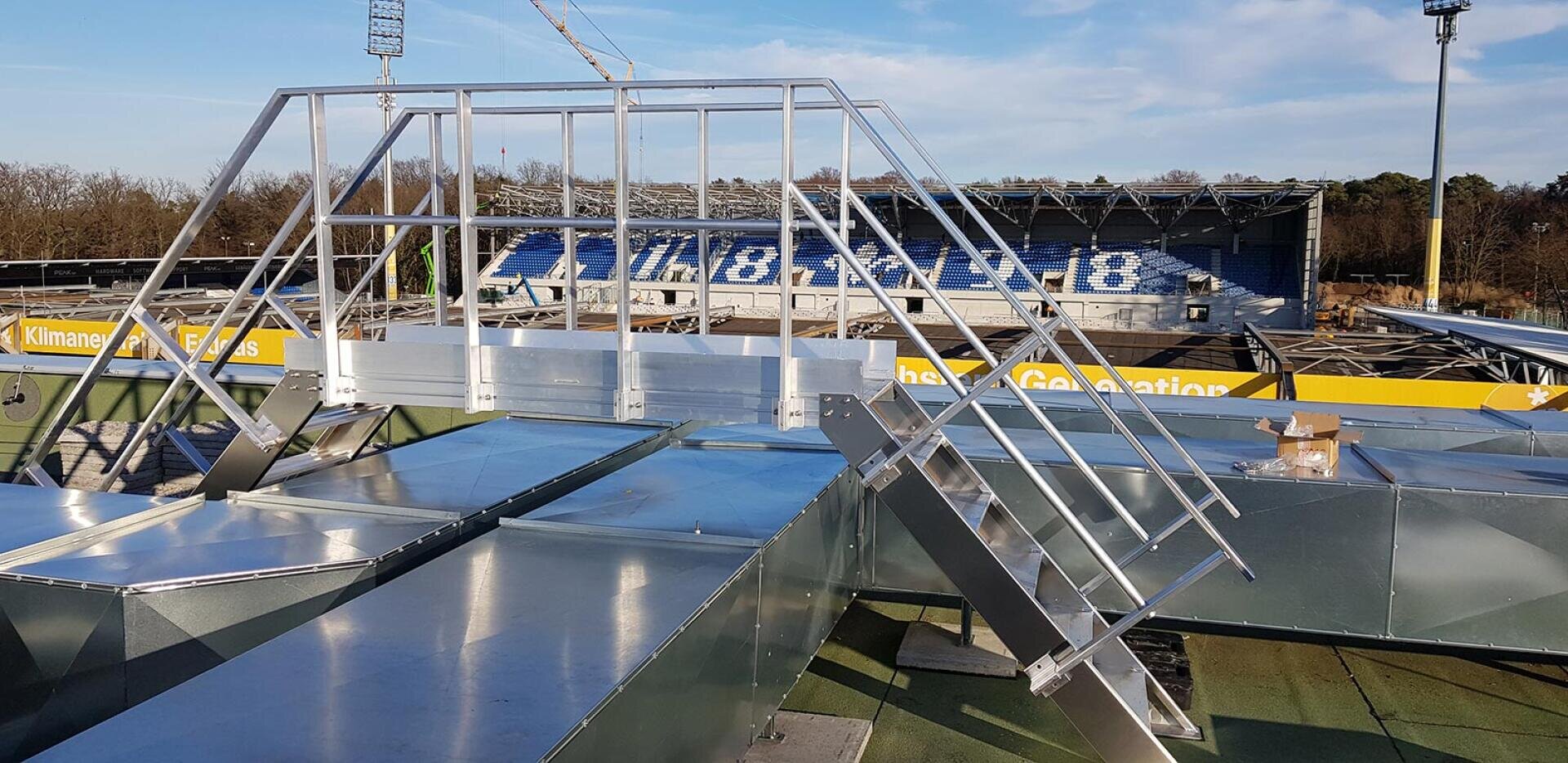 A metal roof walkway with railings and stairs is installed above air shafts and offers a view of a sports stadium with blue seats and yellow advertising. Trees and floodlights can be seen in the background.