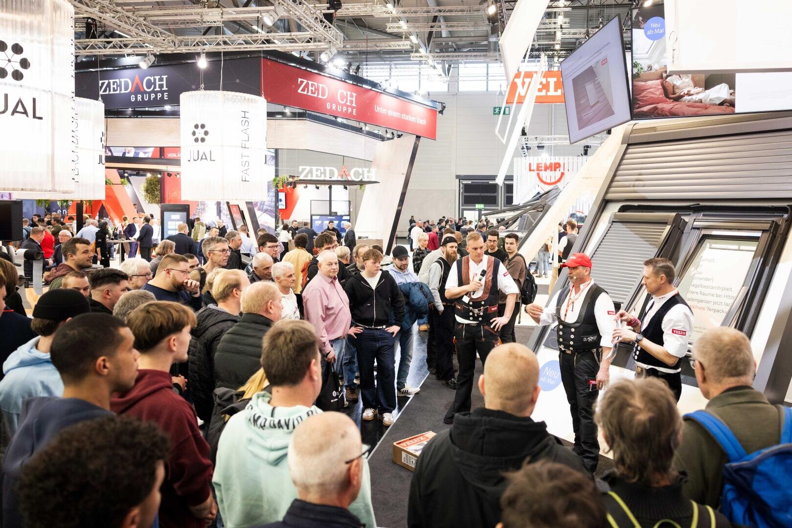 Eine große Gruppe von Menschen hört drei Moderatoren in traditioneller Arbeitskleidung zu, die an einem belebten Messestand in einer Halle, umgeben von Ausstellungsständen und Produktdisplays, eine Vorführung geben.