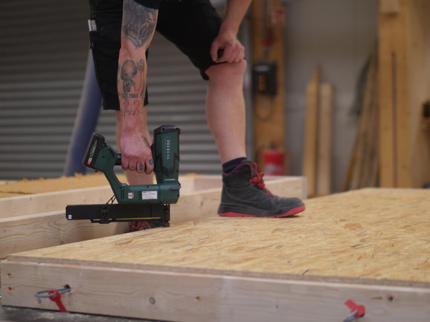 Eine tätowierte Person arbeitet in einer Werkstatt mit einem Elektrowerkzeug, um eine Litzenplatte an einem Holzrahmen zu befestigen. Sie trägt schwarze Shorts und schwarze Schuhe mit roten Schnürsenkeln.