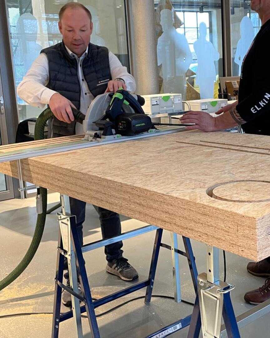 Ein Mann schneidet mit einer Kreissäge ein auf Sägeböcke gestütztes Holzbrett in einer Halle, während eine andere Person das Brett stützt. Um sie herum sind Bauwerkzeuge und -materialien zu sehen.