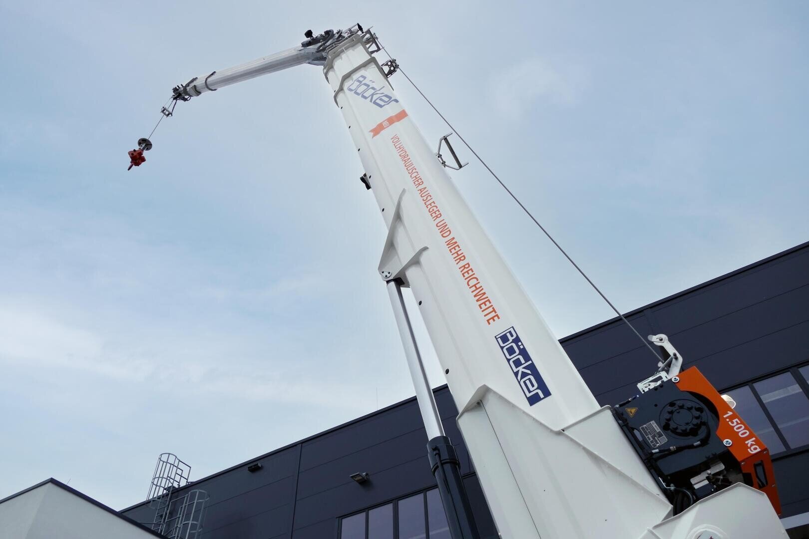 A large white crane with Böcker lettering towers high into the sky next to a modern industrial building, viewed diagonally from below. The crane hook can be seen at the end of the long arm.