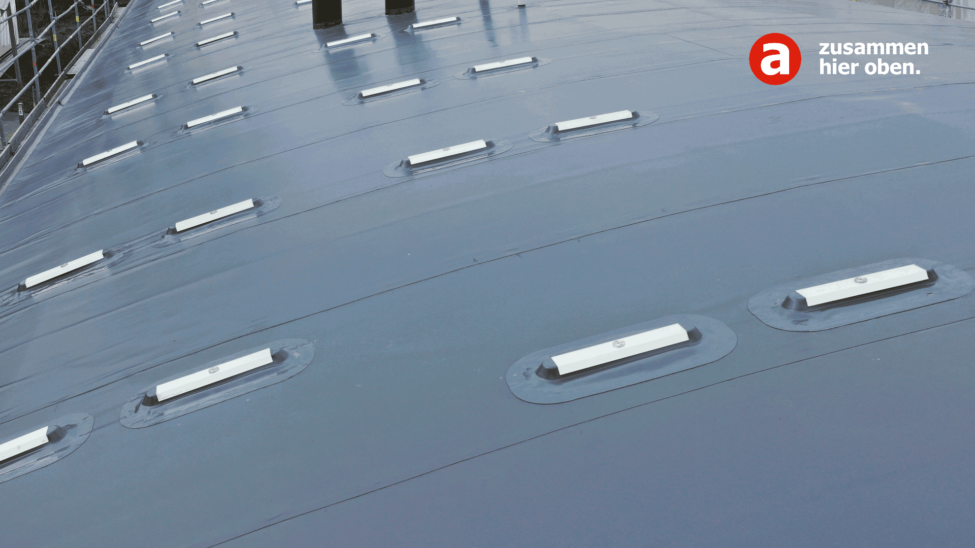 Ein glattes, geneigtes Metalldach mit Reihen rechteckiger Oberlichter. In der oberen rechten Ecke sind das Logo „a“ und der deutsche Text „zusammen hier oben“ sichtbar.