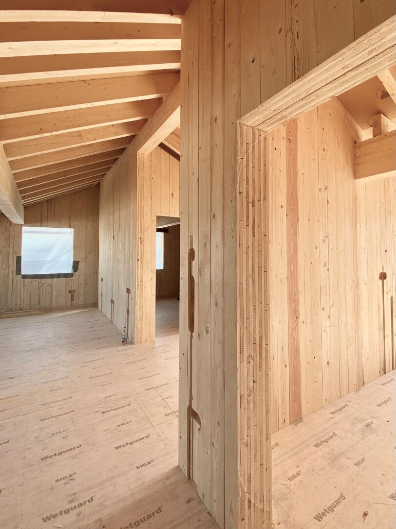 Innenansicht eines im Bau befindlichen Gebäudes mit unfertigen Holzwänden, -decken und -böden. Große Fenster lassen natürliches Licht herein und unterstreichen das geräumige, offene Design und die sichtbaren Balken.