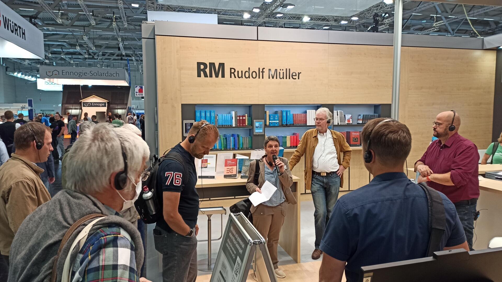 Eine Gruppe von Menschen mit Headsets hört einer Frau zu, die am Stand von Rudolf Müller auf einer Messe, umgeben von Büchern und Ausstellungsmaterialien, in ein Mikrofon spricht.