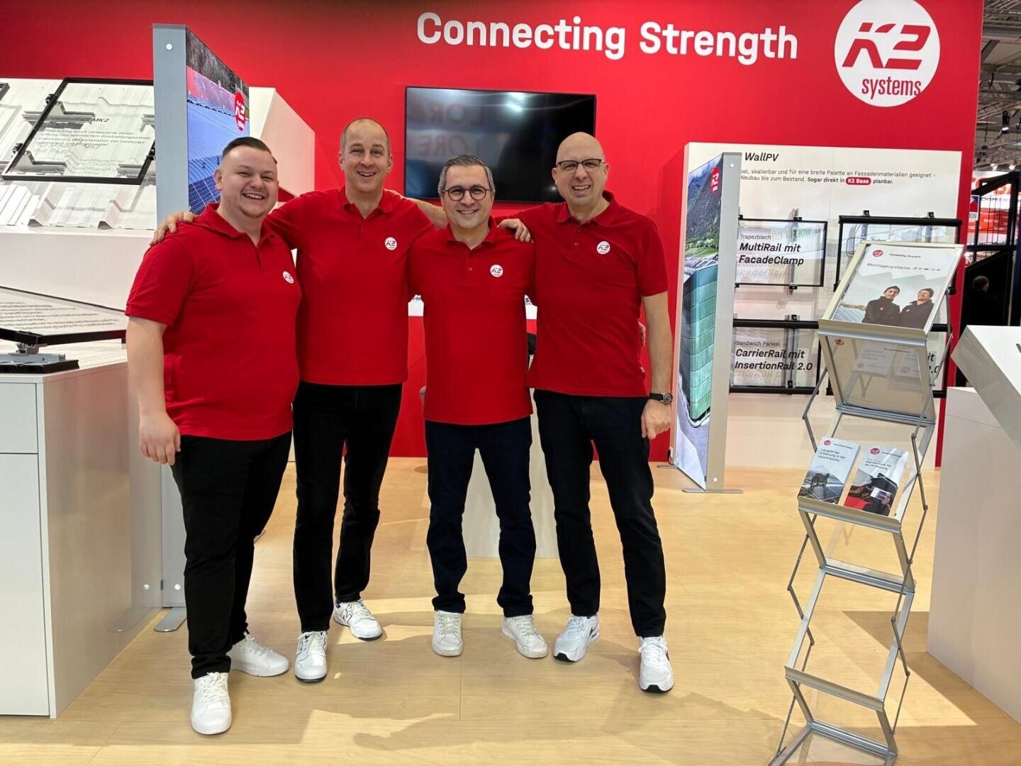 Vier Männer in roten Poloshirts und weißen Turnschuhen von K2 Systems stehen lächelnd vor einem roten Messestand mit der Aufschrift Connecting Strength. Broschüren und Ausstellungsmaterial sind sichtbar.