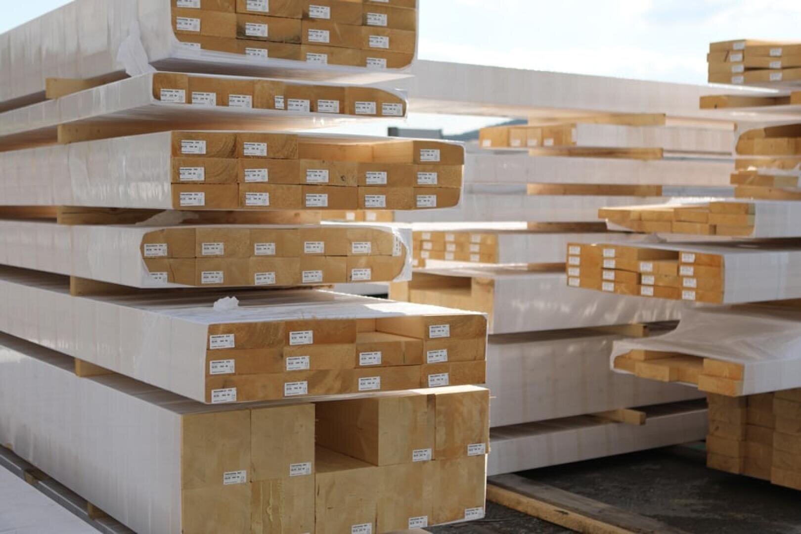Stapel von großen, rechteckigen Holzbalken sind in weißes Plastik eingewickelt und im Freien aufgestellt. Jeder Balken ist mit einem kleinen weißen Etikett versehen, und die Balken sind ordentlich in Reihen übereinander gestapelt.