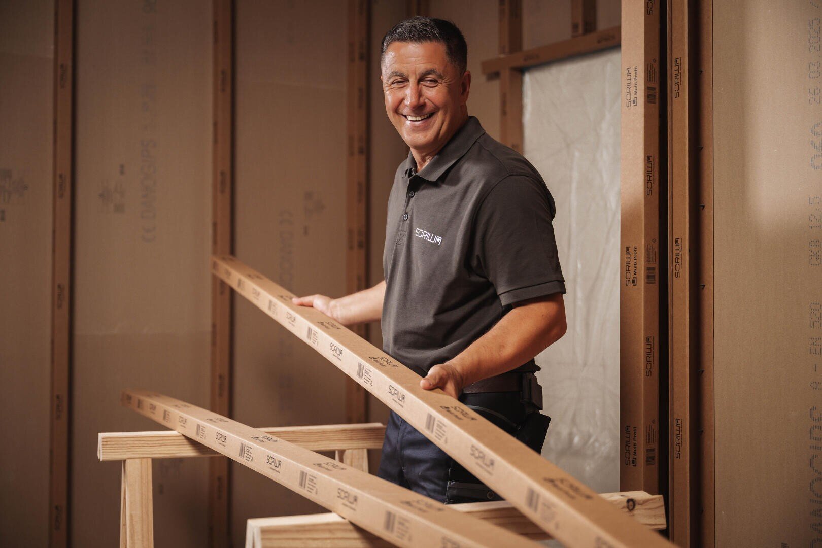 Ein lächelnder Mann in einem grauen Polohemd hält zwei lange Holzbretter in ein im Bau befindliches Gebäude mit freiliegendem Gebälk und Dämmplatten aus Pappe.