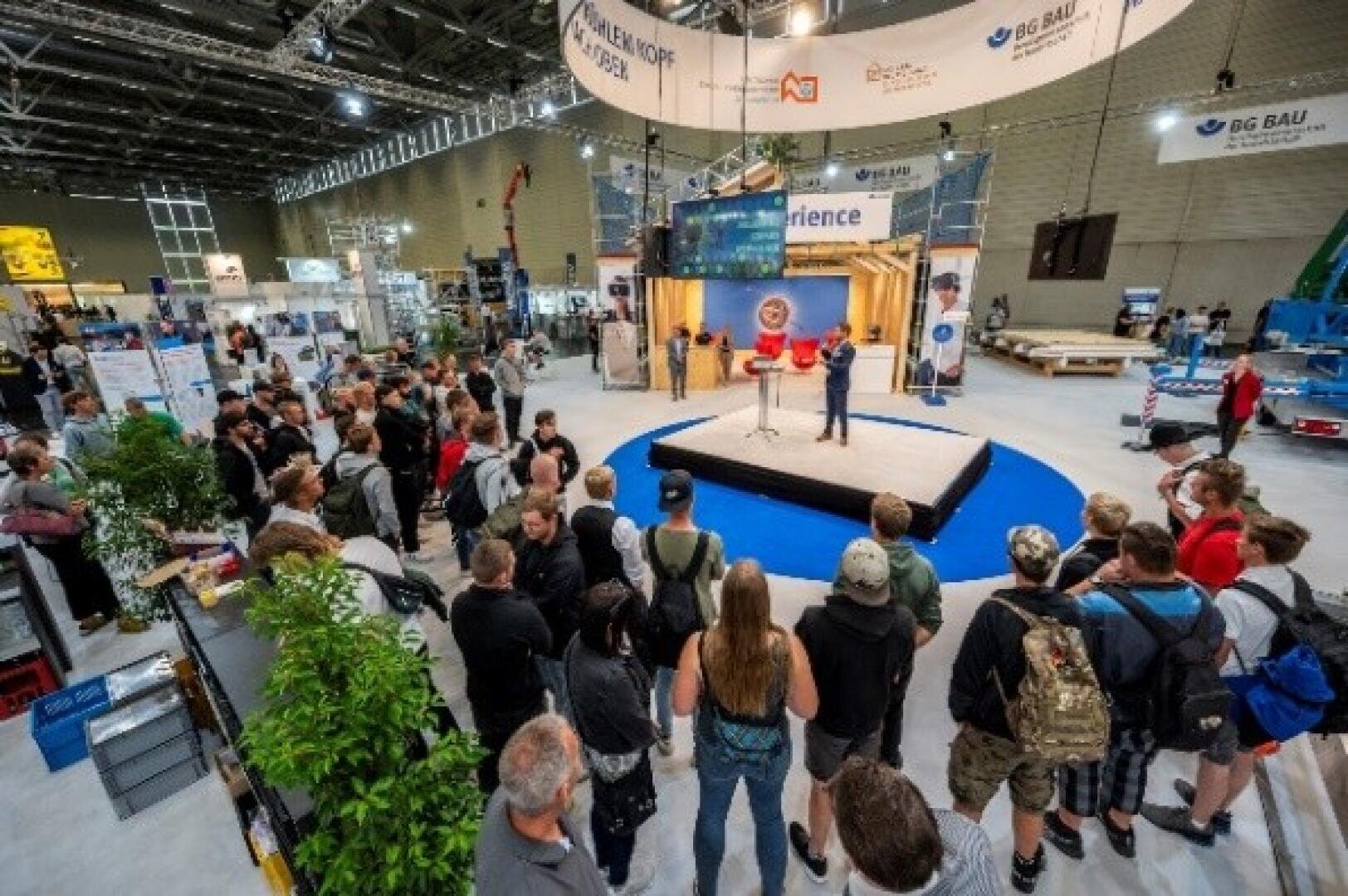 A crowd gathers around a small stage at a covered trade show or expo and listens to a speaker. Various stands, banners and equipment displays can be seen in the spacious, well-lit exhibition hall.