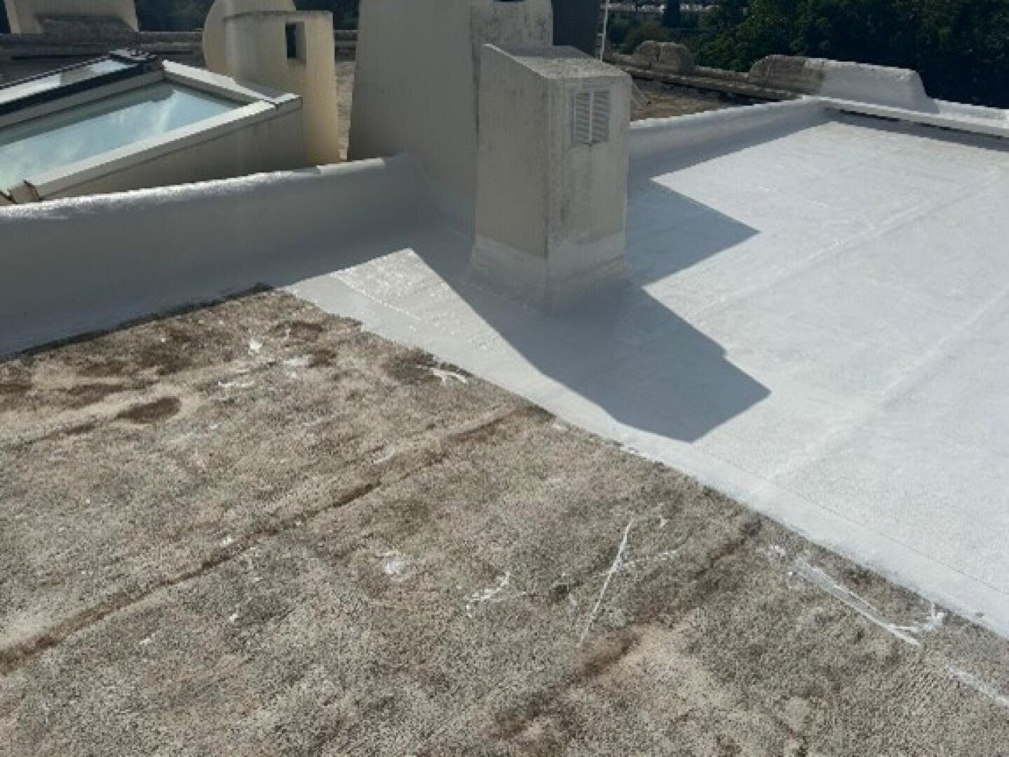 Ein Dach mit einem Abschnitt der alten, verwitterten Oberfläche neben einer neu aufgebrachten weißen, wasserdichten Beschichtung, getrennt durch eine deutliche Linie. Im Hintergrund sind Dachkonstruktionen und ein gläsernes Oberlicht zu sehen.